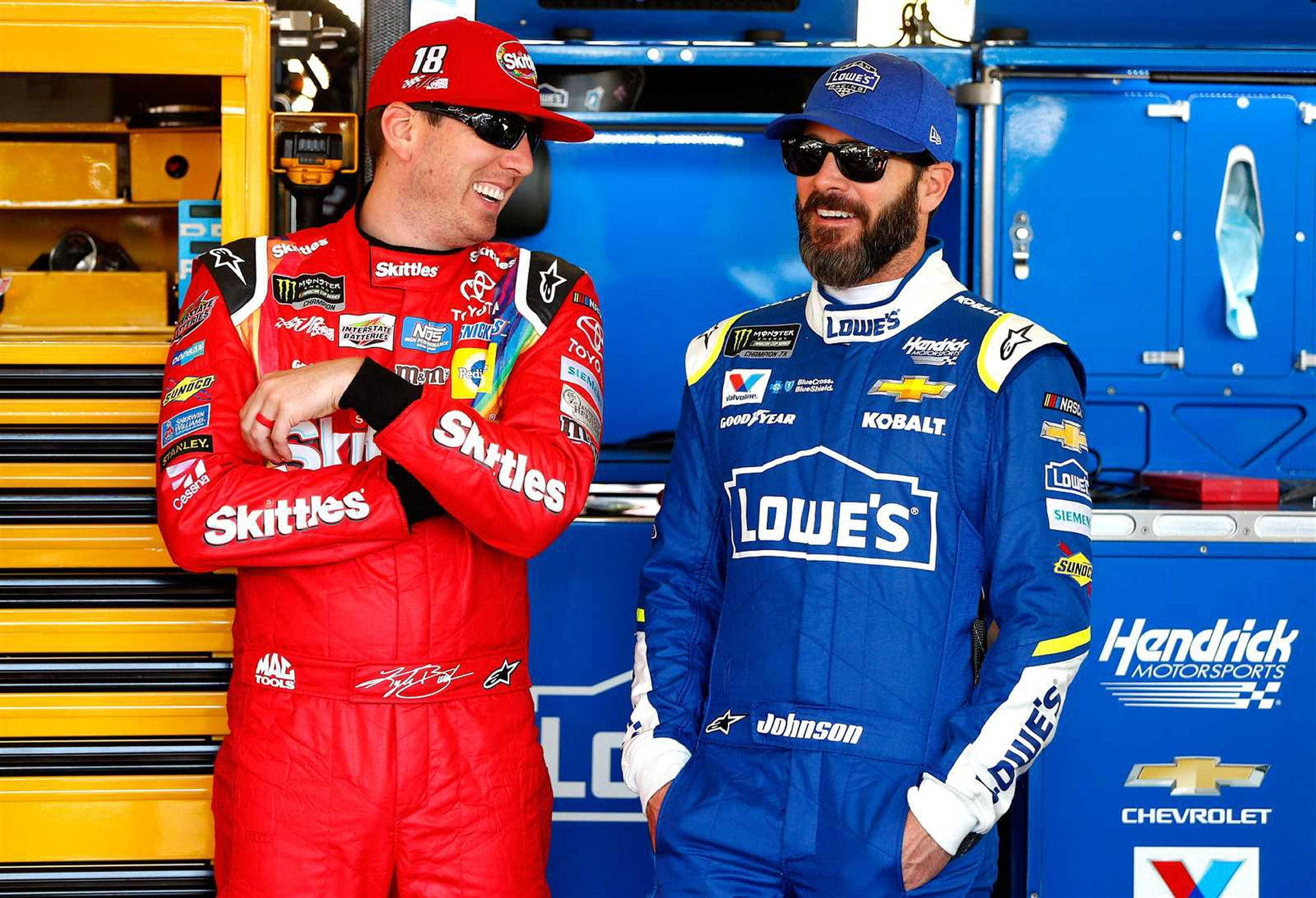 凱爾·布希與吉米·約翰遜 壁紙 — 兩名身穿紅色和藍色賽車服的NASCAR車手在一個有黃色工具箱和藍色設備的車庫裡一起大笑。