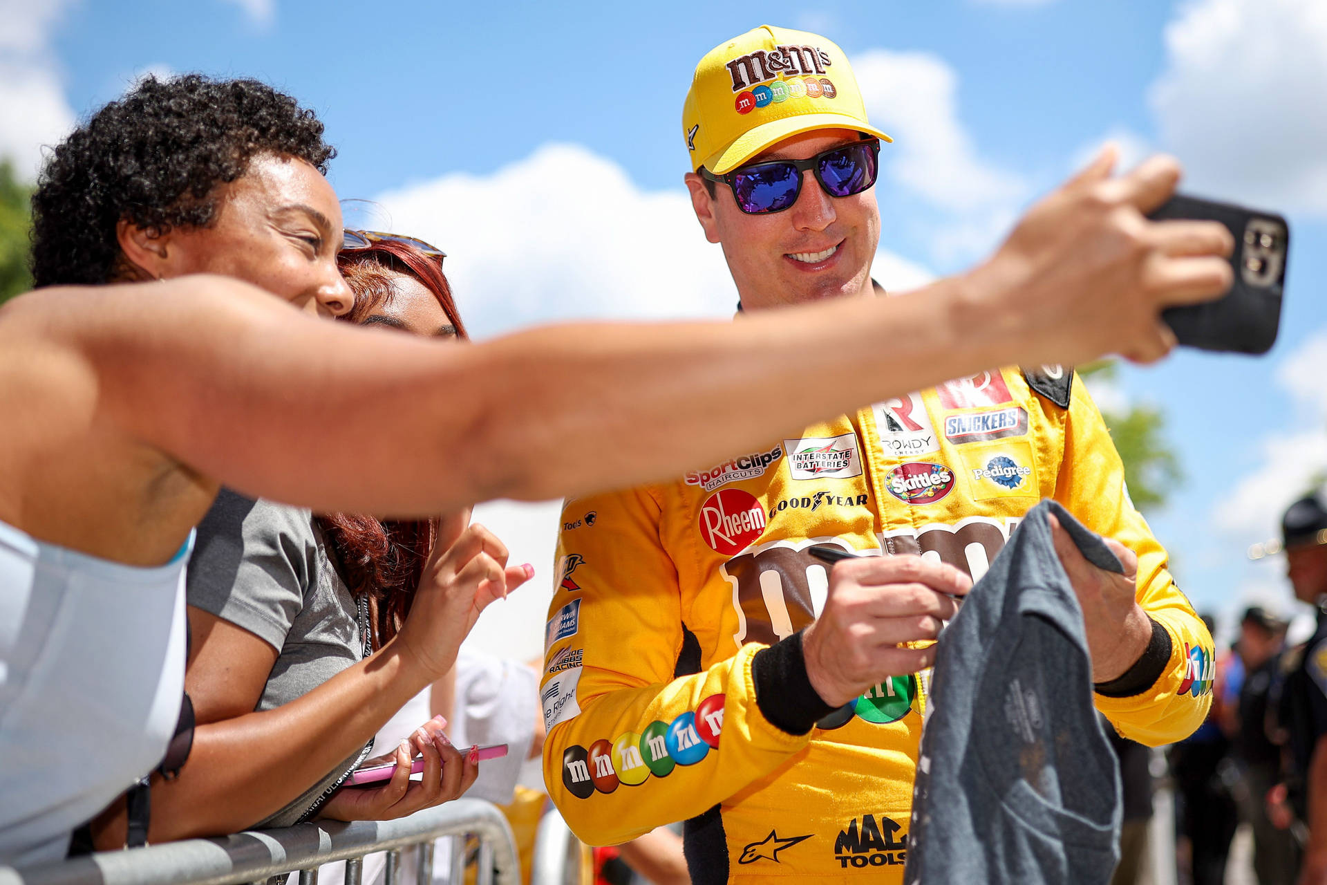 凱爾·布希與粉絲合影 壁紙 — 一位微笑的NASCAR車手穿著鮮黃色M&M's賽車服，在晴朗的藍天下與粉絲自拍。