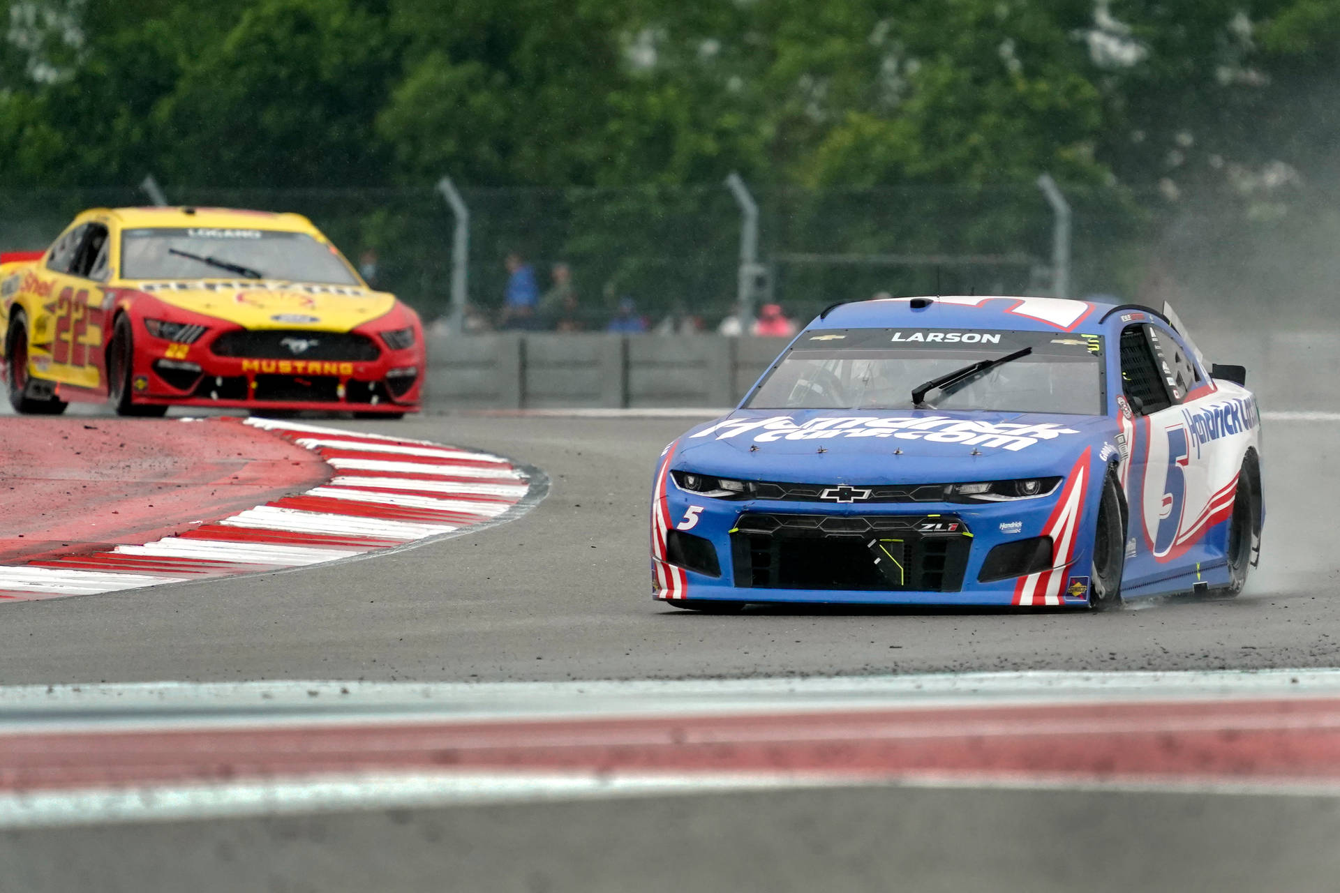 兩輛NASCAR賽車在潮濕的賽道上奔馳，輪胎濺起水花。