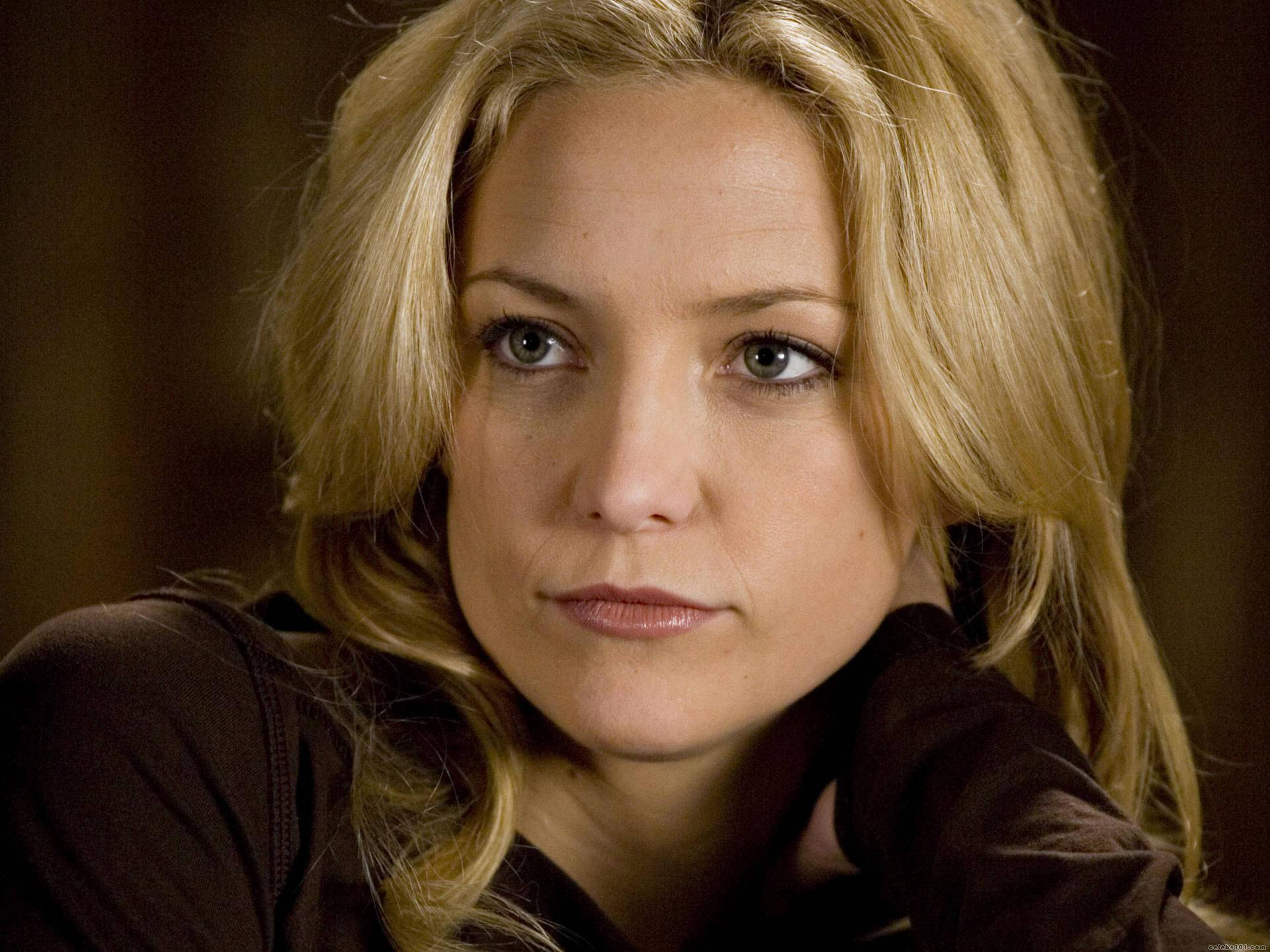 凱特·哈德森隨性姿勢 壁紙 — 一位金髮女性特寫肖像，擁有強烈的藍色眼睛，直視鏡頭，背景為深色模糊。