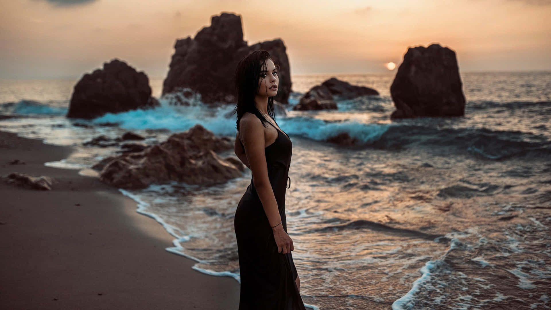 身穿黑色連衣裙的女性在日落時站在海灘上，有海浪和岩石。