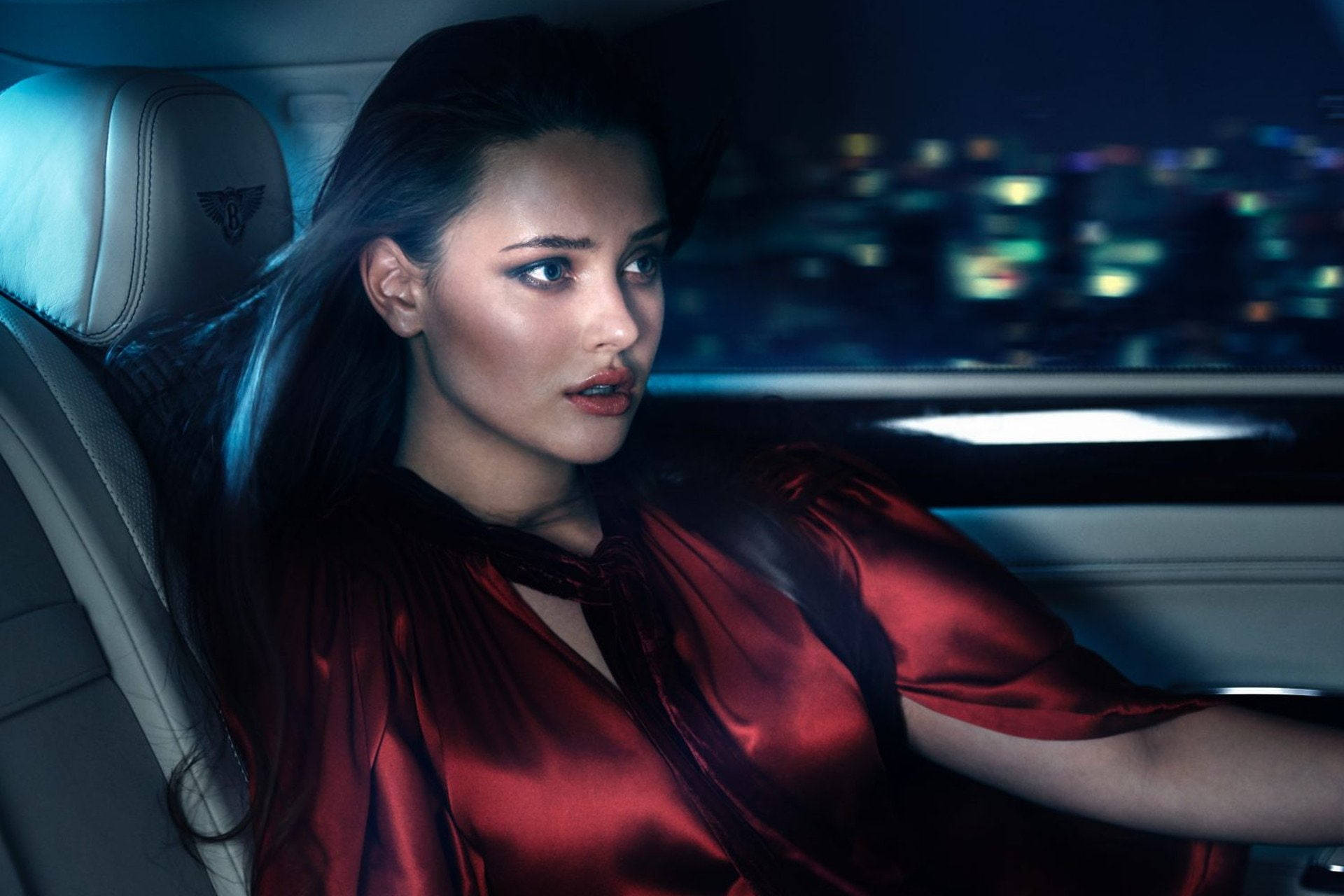 身穿紅色緞面襯衫的女性夜間坐在豪華汽車中，背景是模糊的城市燈光。
