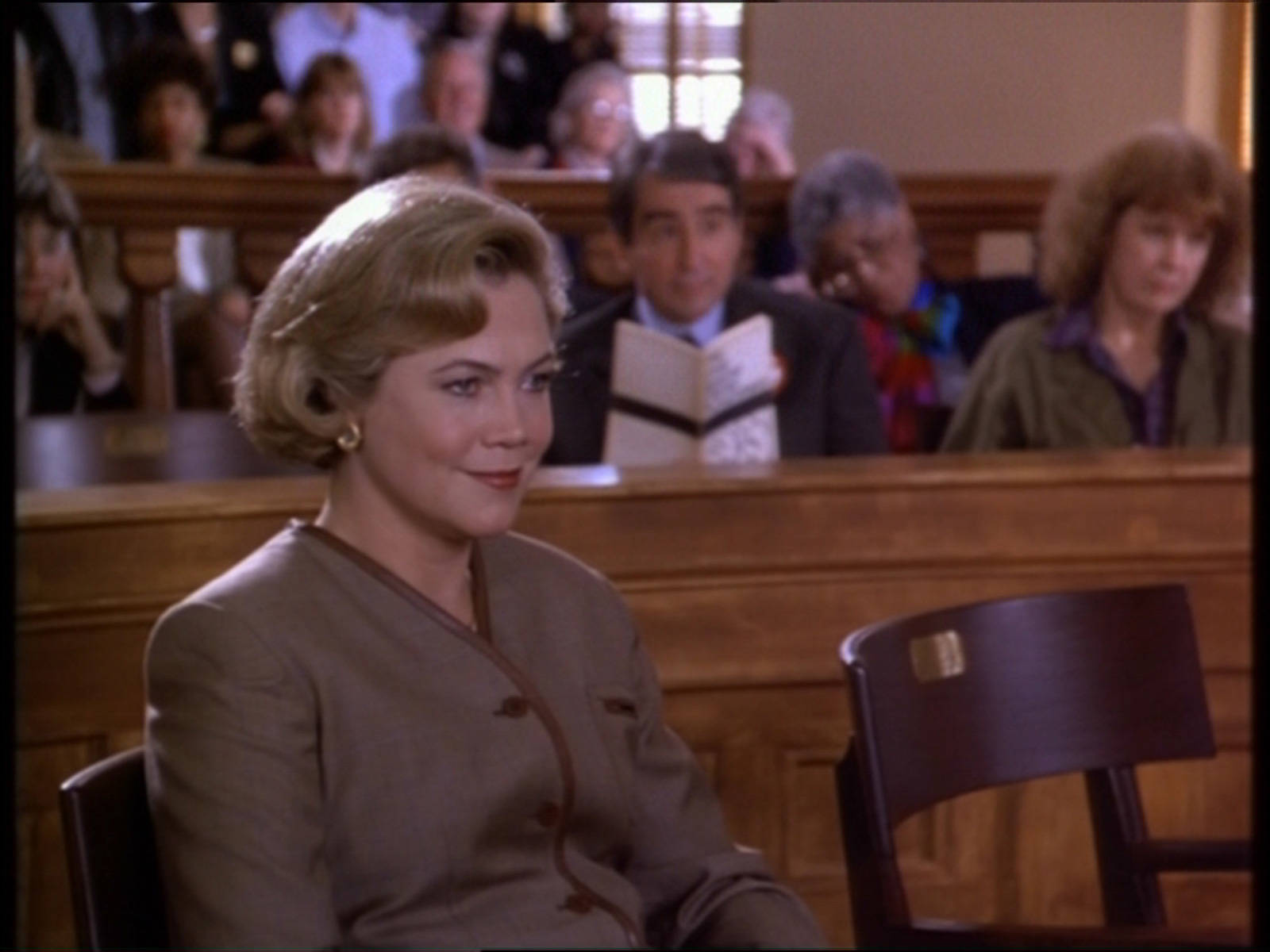 凱瑟琳·特納《Serial Mom》預告片 壁紙 — 一位女性在法庭上坐著，露出微妙的笑容，身後木製長椅上坐著模糊的旁觀者。