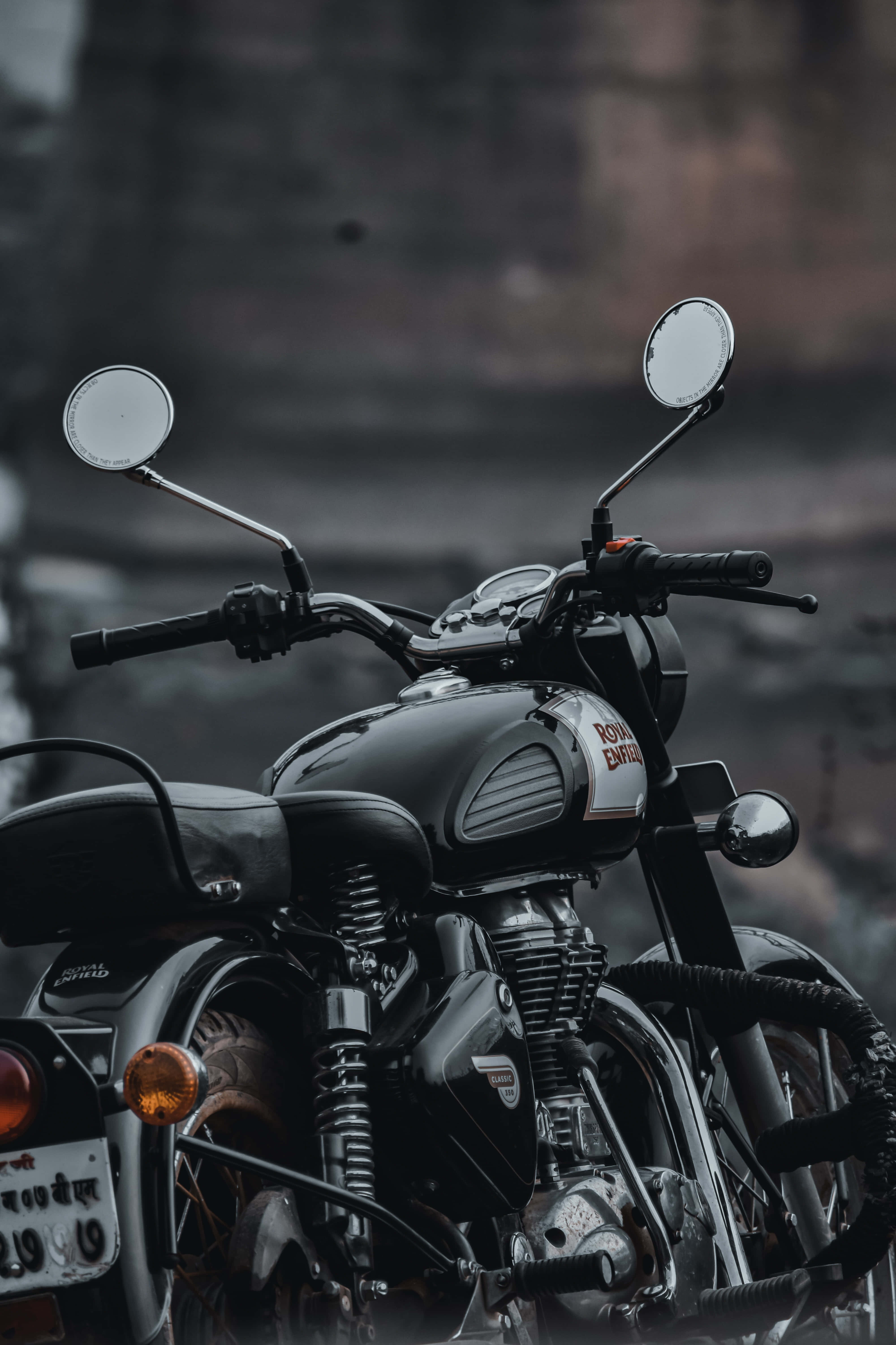 出色的黑色皇家恩菲爾德機車背景 壁紙 — 一輛時尚的黑色 Royal Enfield 電單車，帶有鍍鉻裝飾和圓形後視鏡，在模糊的城市背景下蓄勢待發。