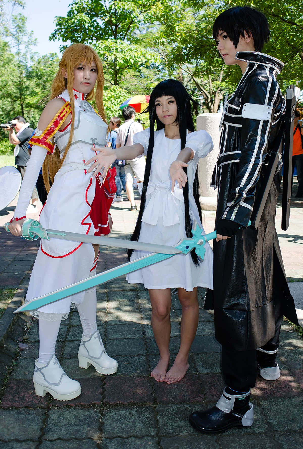 《刀劍神域》cosplay：桐人與亞絲娜 壁紙 — 三位穿著精細動漫服裝的角色扮演者在戶外陽光下，共同手持一把發光的劍擺姿勢。