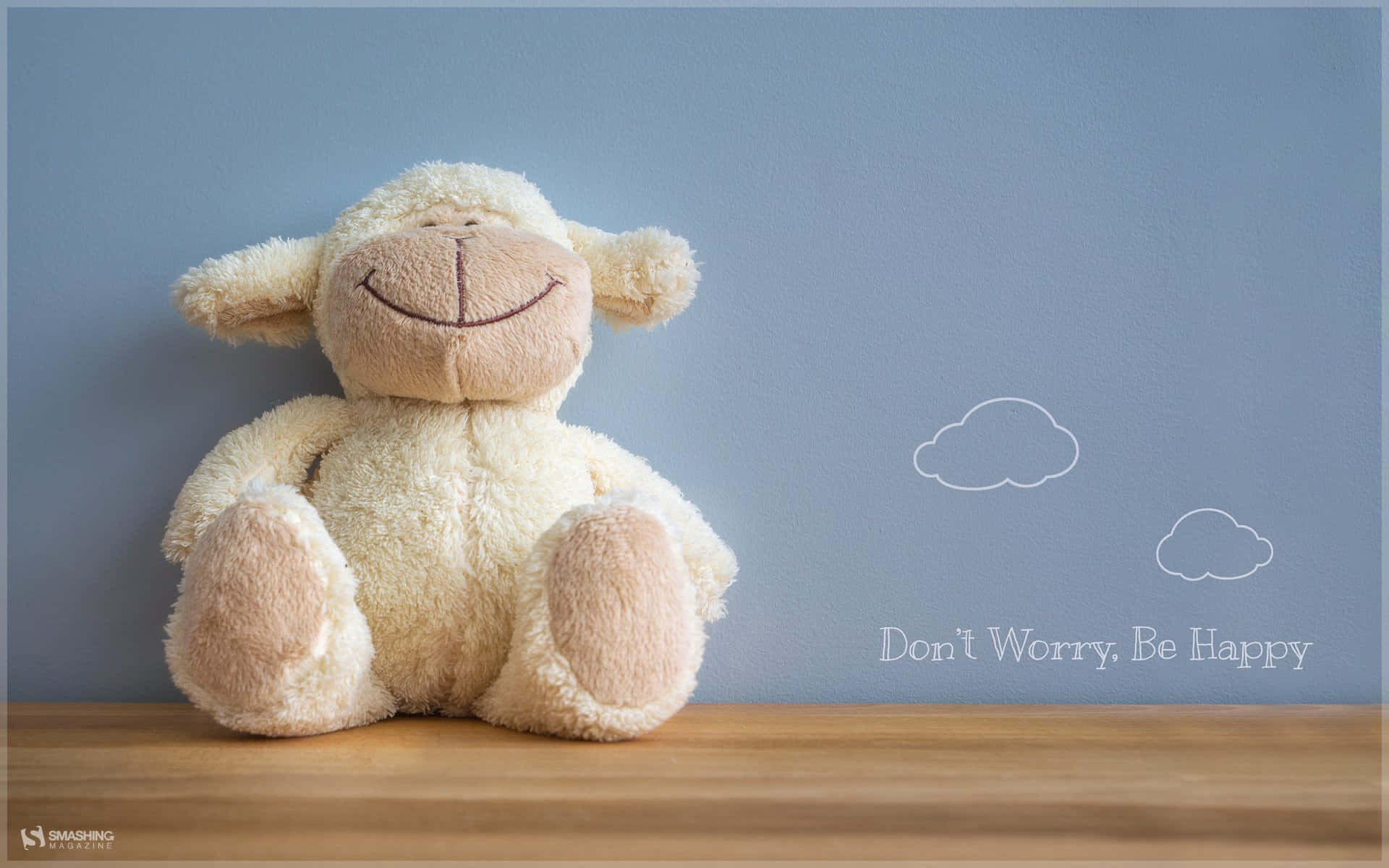 一隻微笑的填充羊在木架上，藍色牆壁上有「Don't Worry. Be Happy」文字和雲朵貼紙。