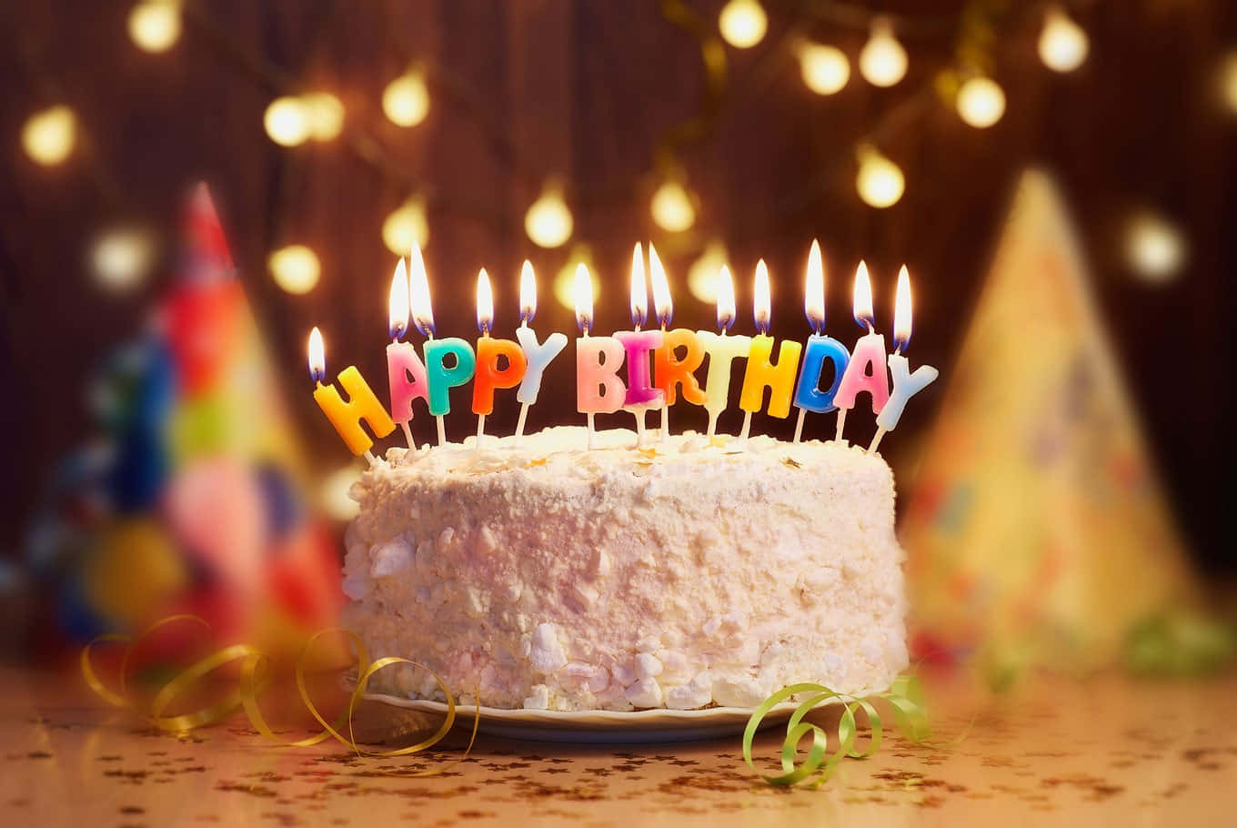分享喜悅：朋友慶祝特別時刻 壁紙 — 一個糖霜生日蛋糕，上面有色彩繽紛的「HAPPY BIRTHDAY」蠟燭，在溫暖的燈串和派對帽背景下閃爍。