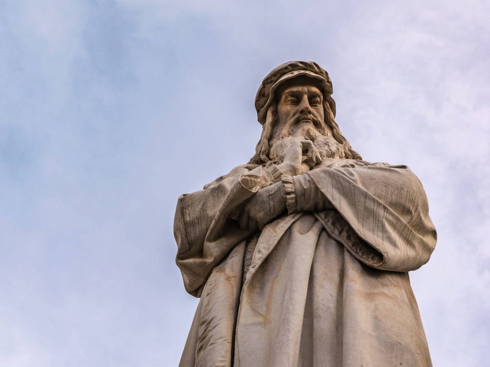 列奧納多·達文西米蘭雕像 壁紙 — 一座戴帽穿袍的鬍鬚男人石像聳立在淡藍色天空和柔和雲朵前。