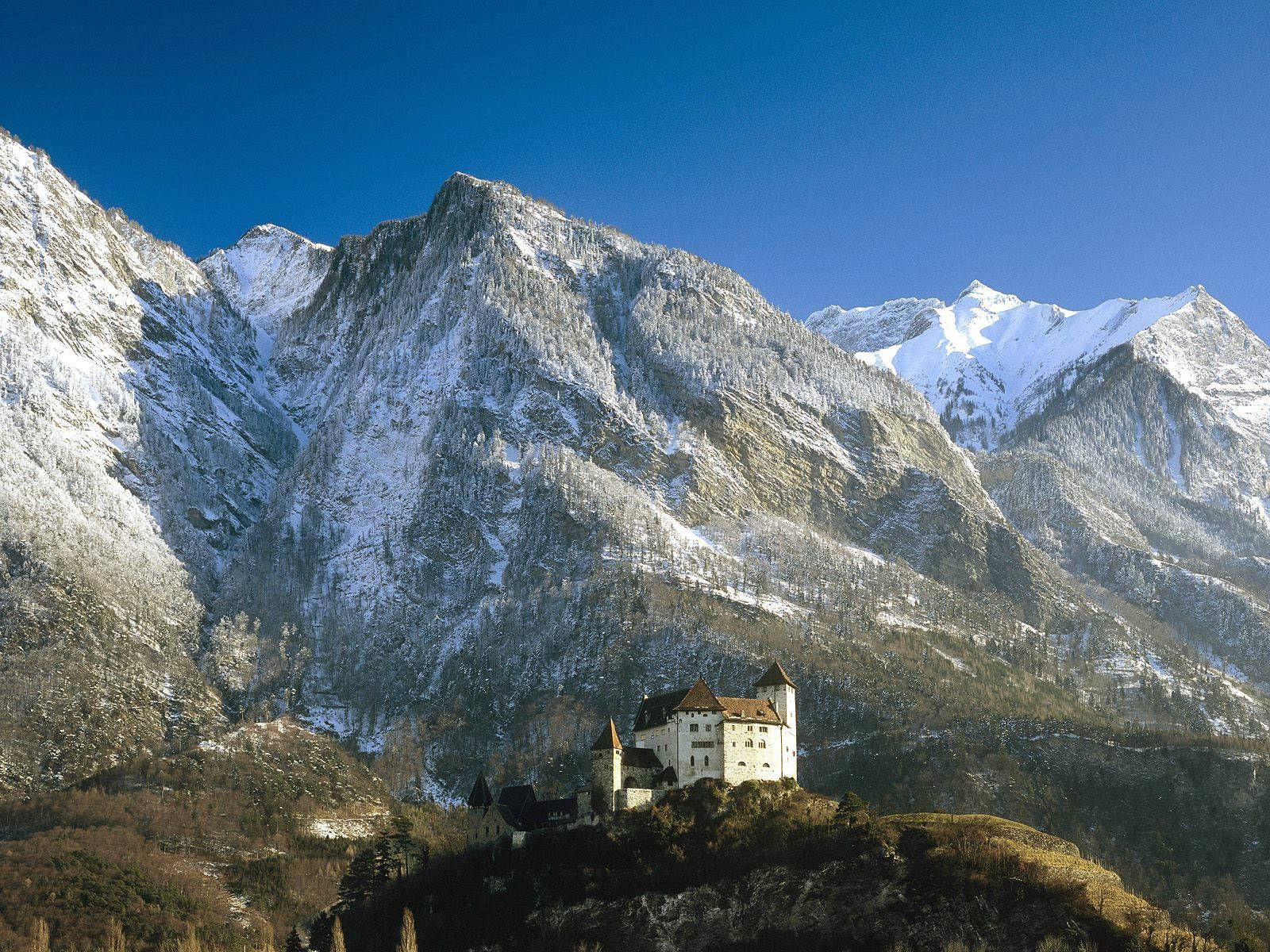 列支敦士登古騰堡城堡 壁紙 — 一座雄偉的雪山山脈聳立在一個歷史悠久的城堡之上，城堡坐落在山坡上，背景是清澈的藍天。