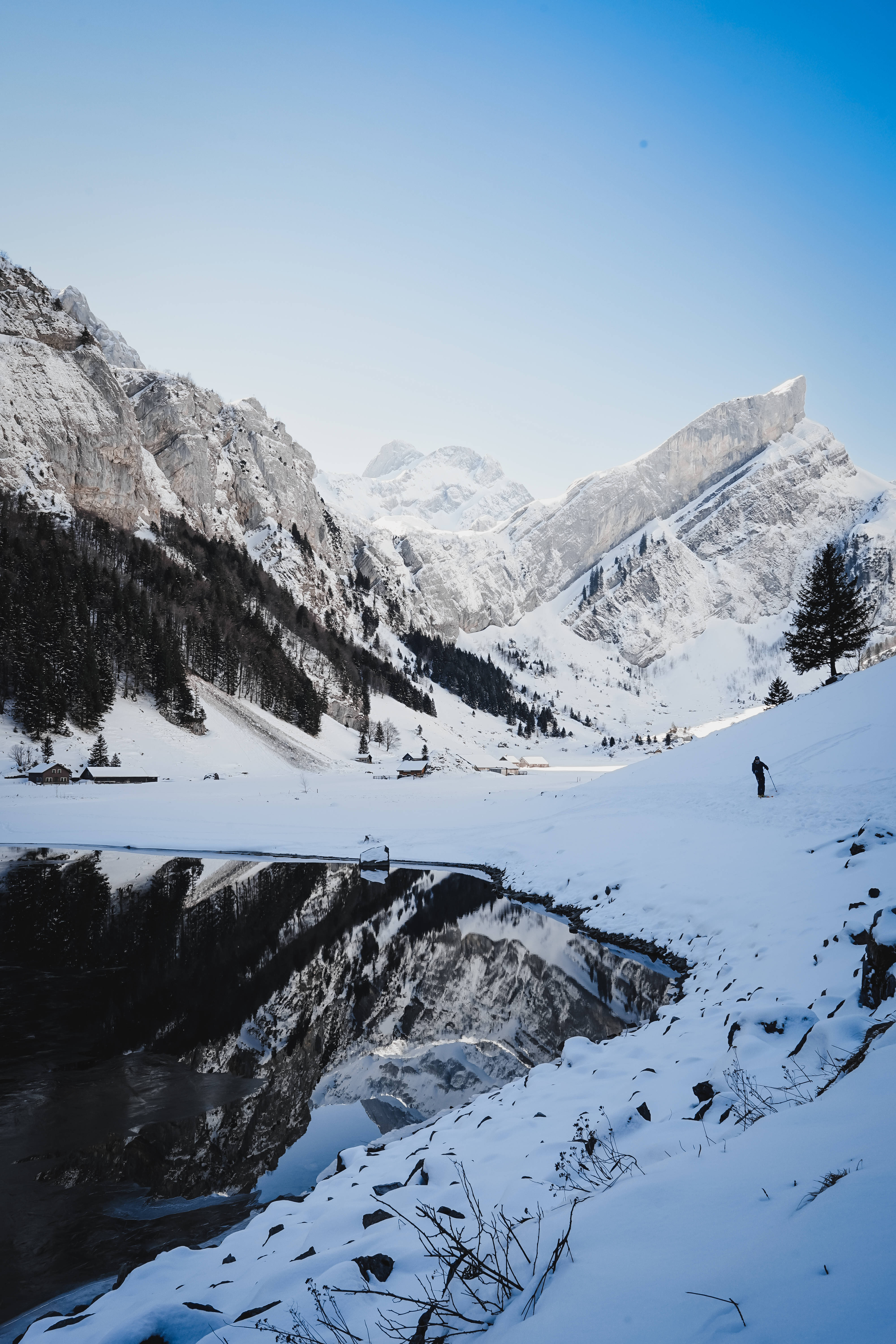 列支敦士登雪山 壁紙 — 一名孤獨的滑雪者正在白雪覆蓋的斜坡上滑下，旁邊是一個結冰的湖泊，湖面映照著高聳的阿爾卑斯山峰，天空清澈湛藍。