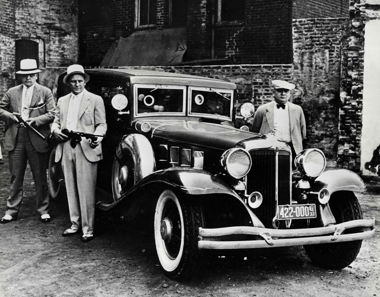 三名身穿西裝和帽子的男子站在一輛1930年代復古汽車旁，背景是磚砌建築。