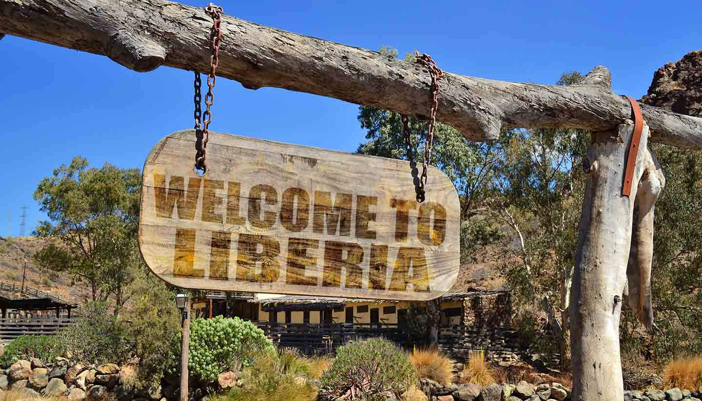 利比里亞木製標誌 壁紙 — 一塊風化的木牌，上面寫著「WELCOME TO LIBERIA」，掛在一個質樸的木框上，背景是乾燥的山丘和藍天。