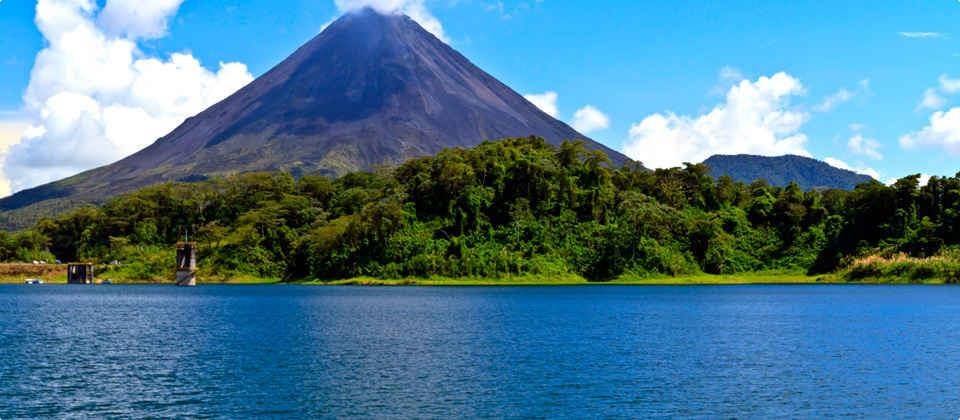 利比里亞藍海山景 壁紙 — 一座雄偉的火山聳立在茂密的綠色森林後面，森林毗鄰平靜的藍色湖泊，上方是明亮的藍天和蓬鬆的白雲。