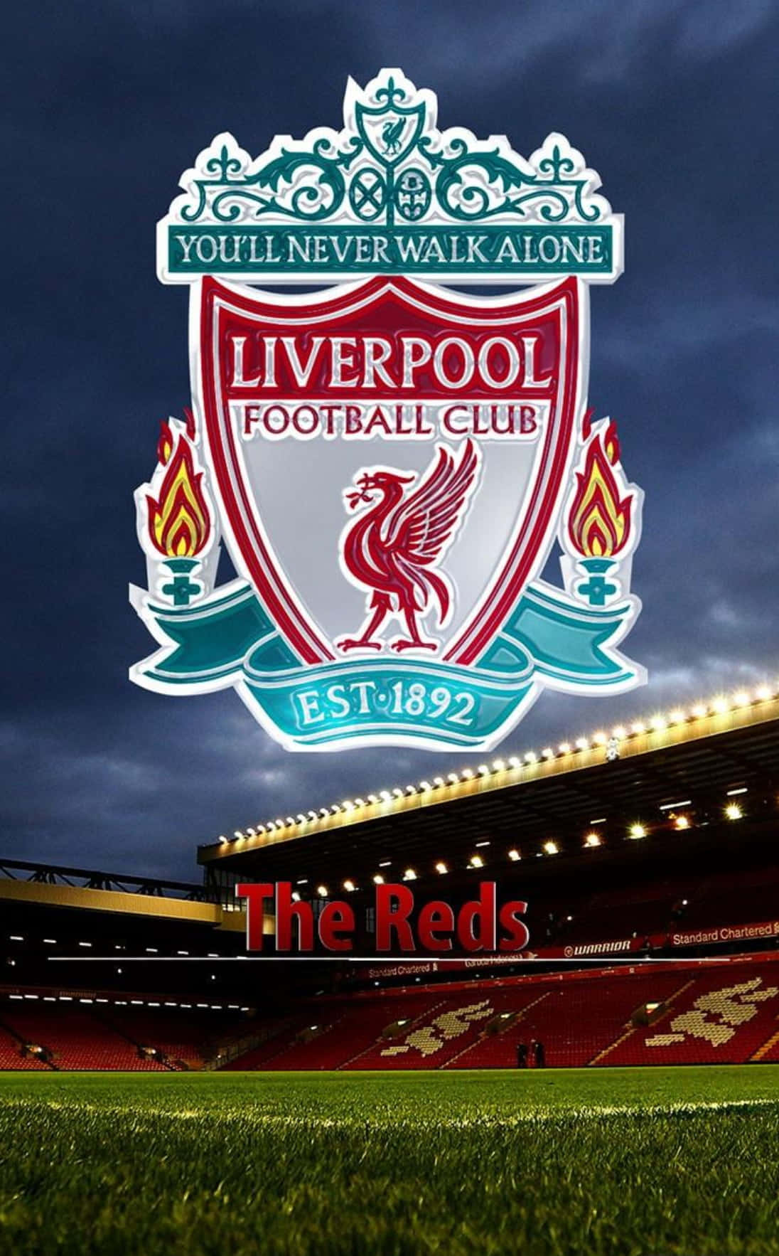 Liverpool Logo 桌布 壁紙 — 利物浦足球會徽章在黑暗天空下，於晏菲路球場上方發光，下方是鮮紅色的座位和綠色草坪。