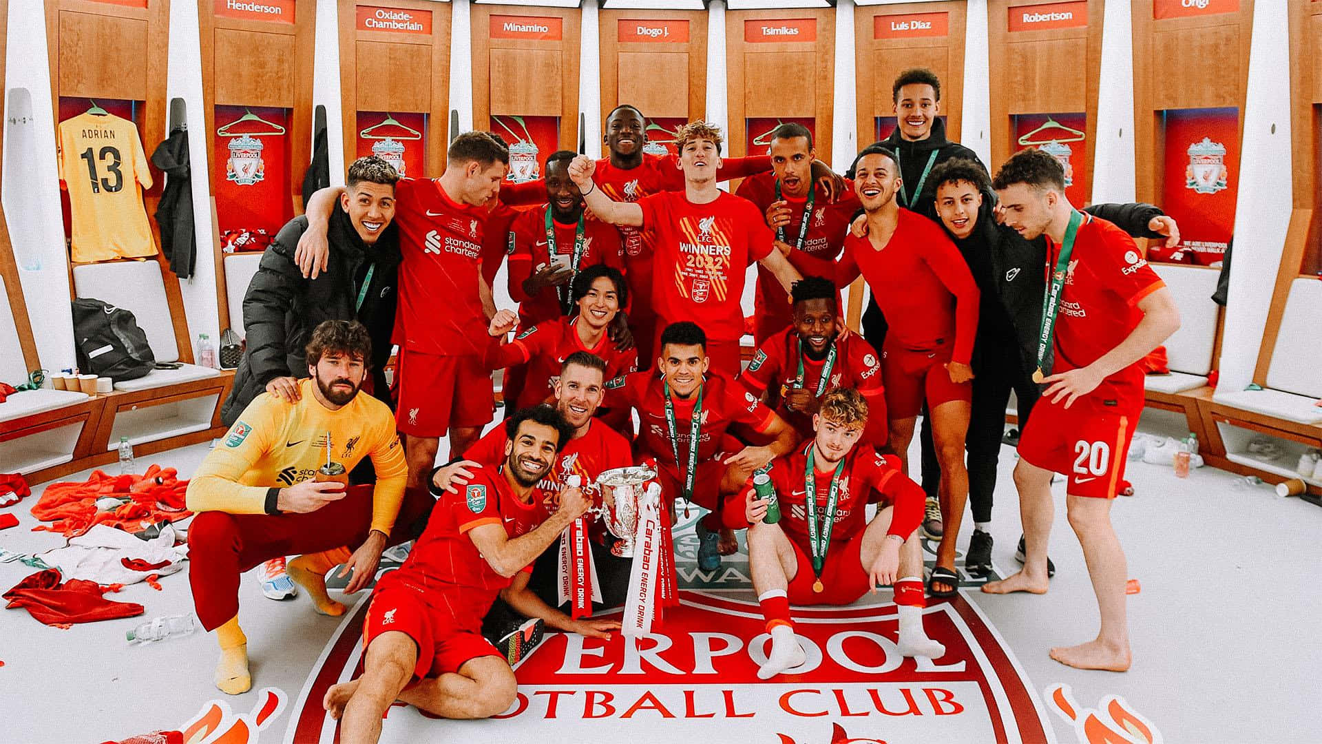 Liverpool 桌布 壁紙 — 利物浦足球隊在更衣室慶祝勝利，手持獎盃和獎牌，周圍是紅色球衣和散落的裝備。