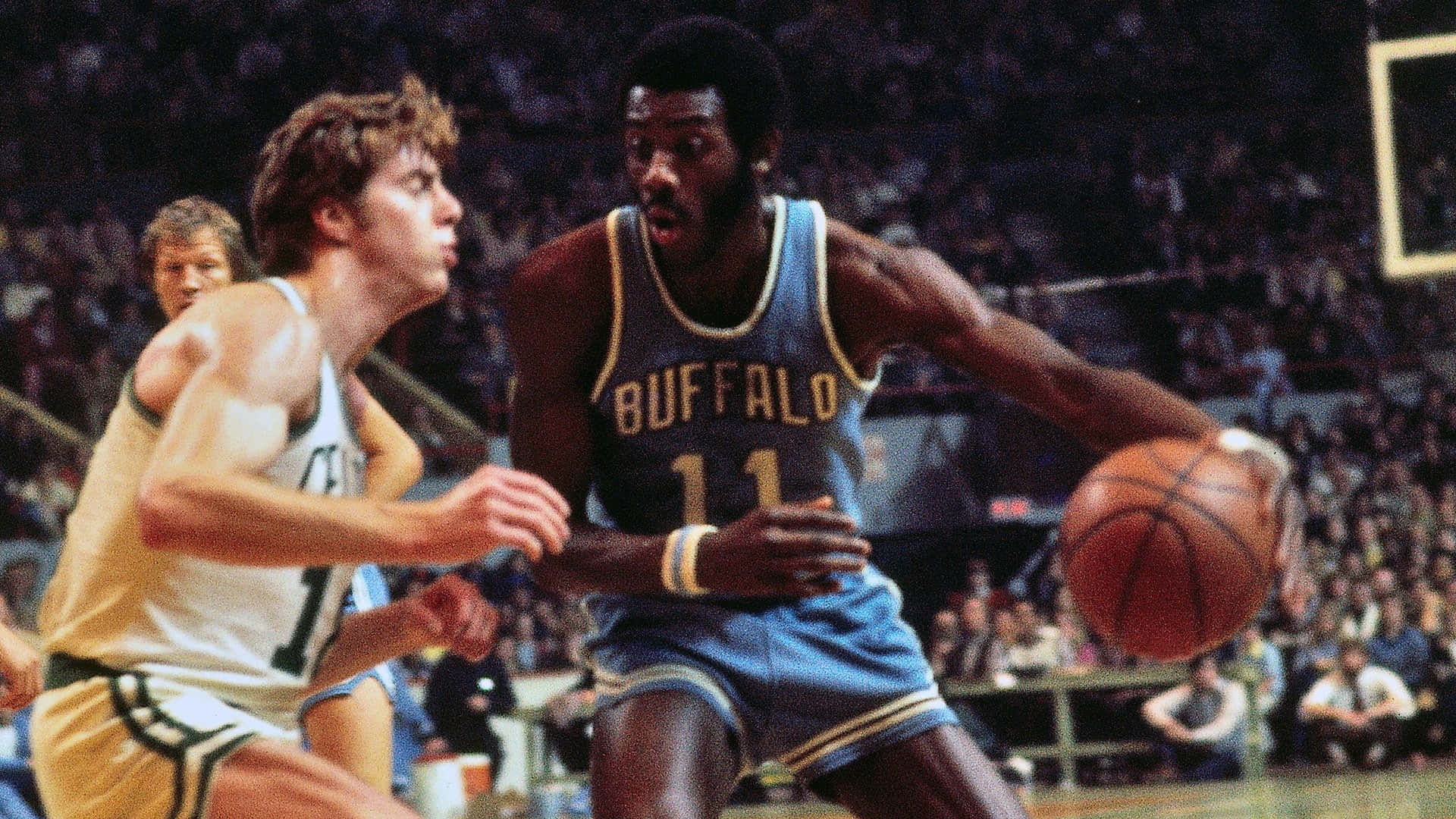 前NBA布法羅勇士隊球員鮑勃·麥卡杜對決戴夫·考文斯 壁紙 — 一名身穿藍色Buffalo球衣的籃球運動員在擠滿觀眾的室內場館中運球，同時被對手防守。