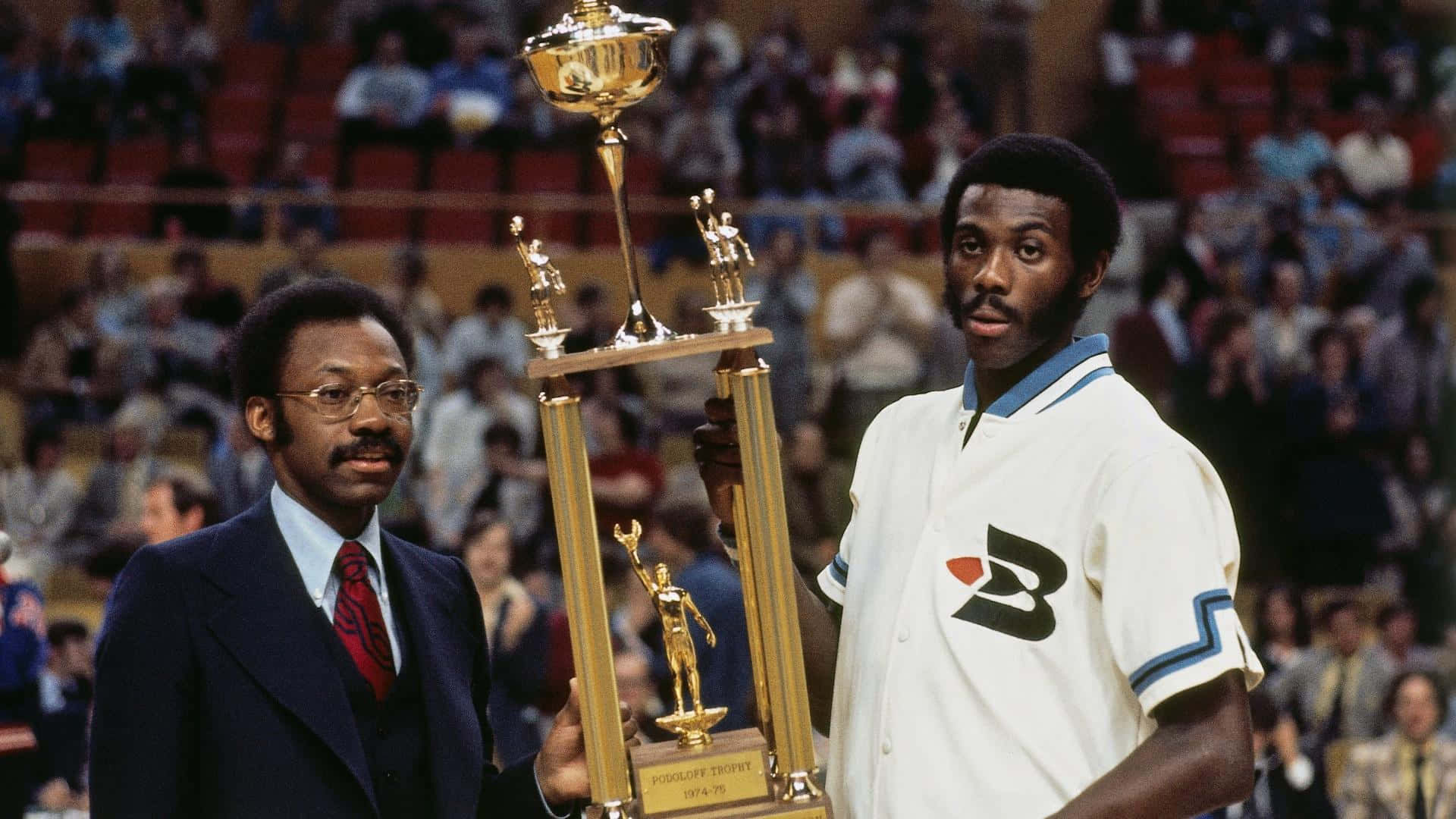 前NBA球員鮑勃·麥卡杜1975年MVP頒獎照 壁紙 — 兩個男人站在擠滿人的場館裡，手持一個大型金色獎盃，獎盃底座上有雕像。
