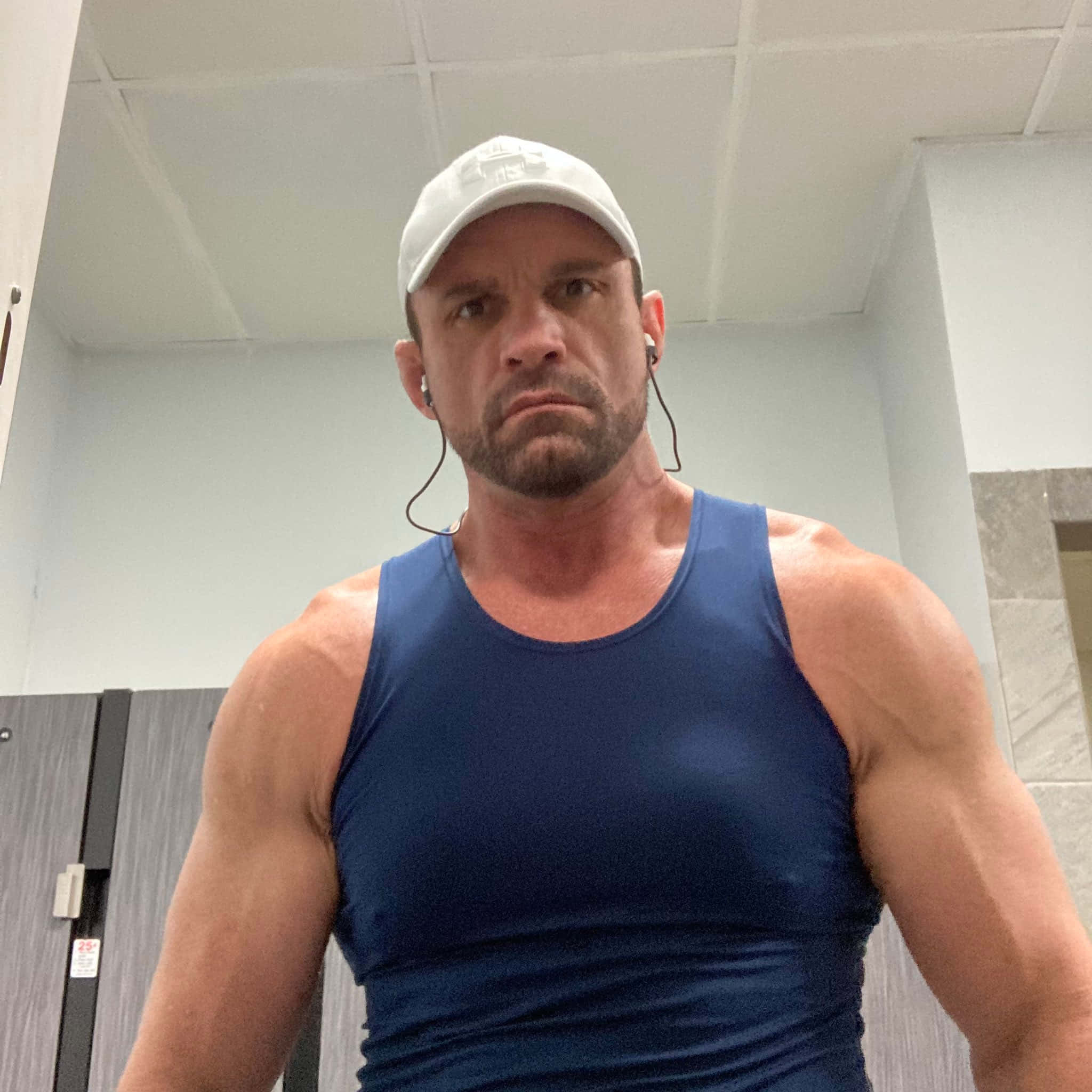 前WWE摔角明星查理·哈斯 壁紙 — 一個肌肉發達的男人穿著藍色背心和白色帽子，在健身室更衣室裡凝視著鏡頭。
