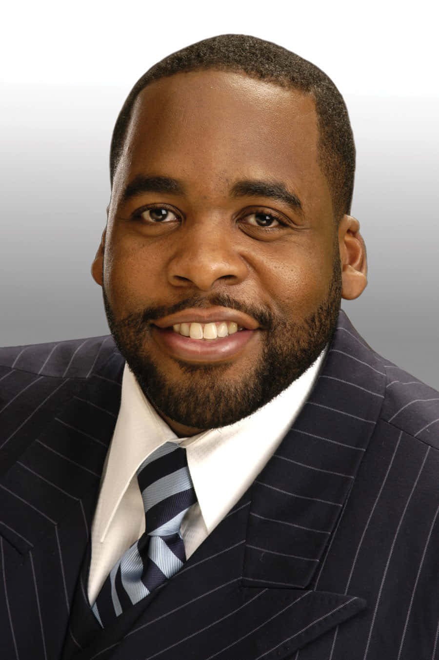 前底特律市長Kwame Kilpatrick肖像 壁紙 — 一位微笑的男子，身穿深色細條紋西裝和條紋領帶，背景為柔和的灰色漸變。