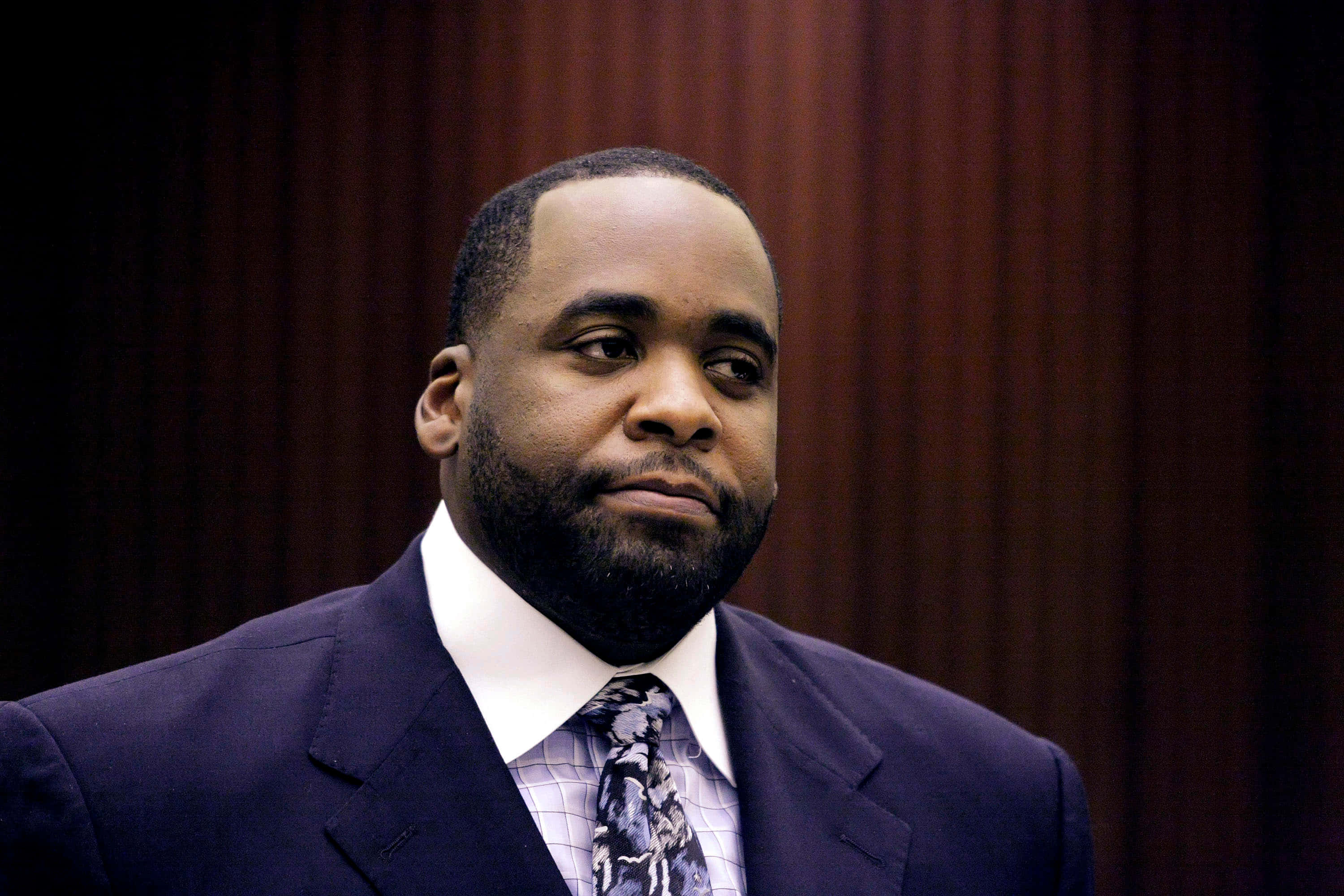 前底特律市長Kwame Kilpatrick沉思時刻 壁紙 — 一名身穿深色西裝和圖案領帶的男子，莊嚴地站在一面豐富的棕色木鑲板牆前。
