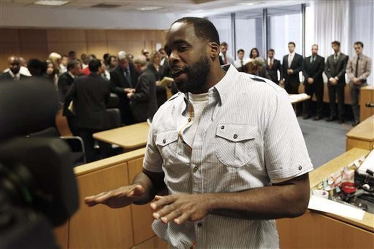 前底特律市長Kwame Kilpatrick出席記者會 壁紙 — 一名身穿條紋襯衫的男子在法庭上說話時做手勢，現場有旁聽者和攝像機。