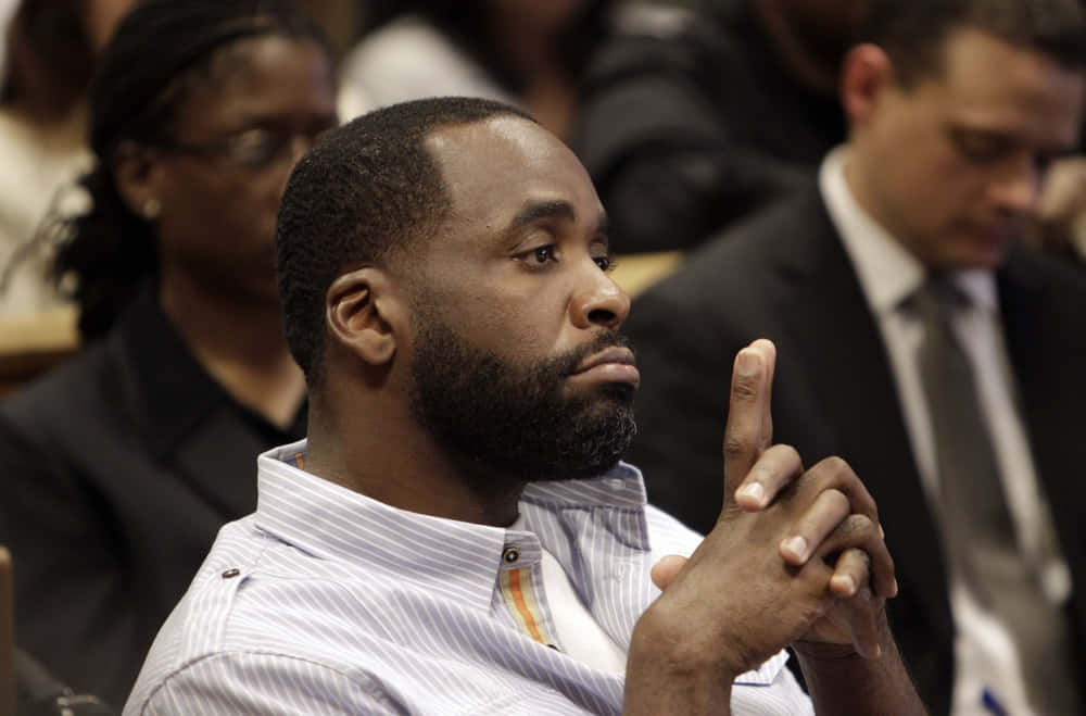 前底特律市長Kwame Kilpatrick出席公開活動 壁紙 — 一個留鬍鬚的男人坐在法庭上，舉起一隻手指，若有所思地望向旁邊。