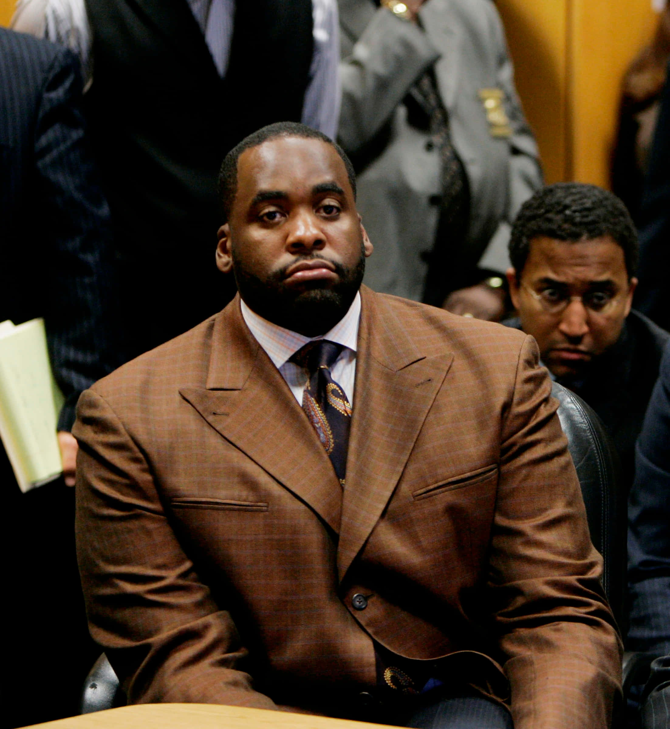 前底特律市長Kwame Kilpatrick 壁紙 — 一名身穿棕色格紋西裝的男子莊嚴地坐在法庭上，周圍的人表情嚴肅。