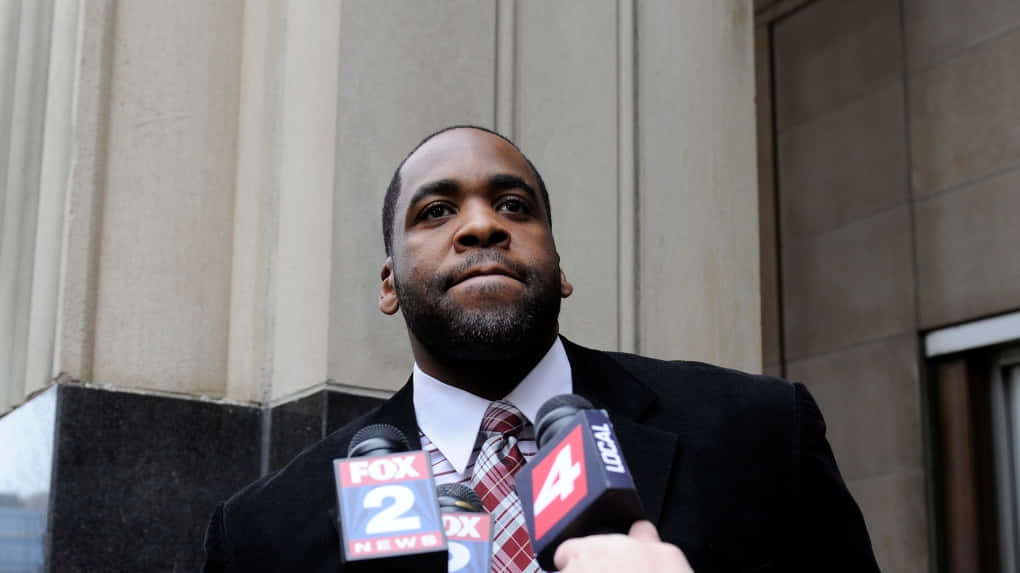 前底特律市長Kwame Kilpatrick官方肖像 壁紙 — 一個穿著西裝的嚴肅男子站在石柱前，記者們將麥克風對準他。