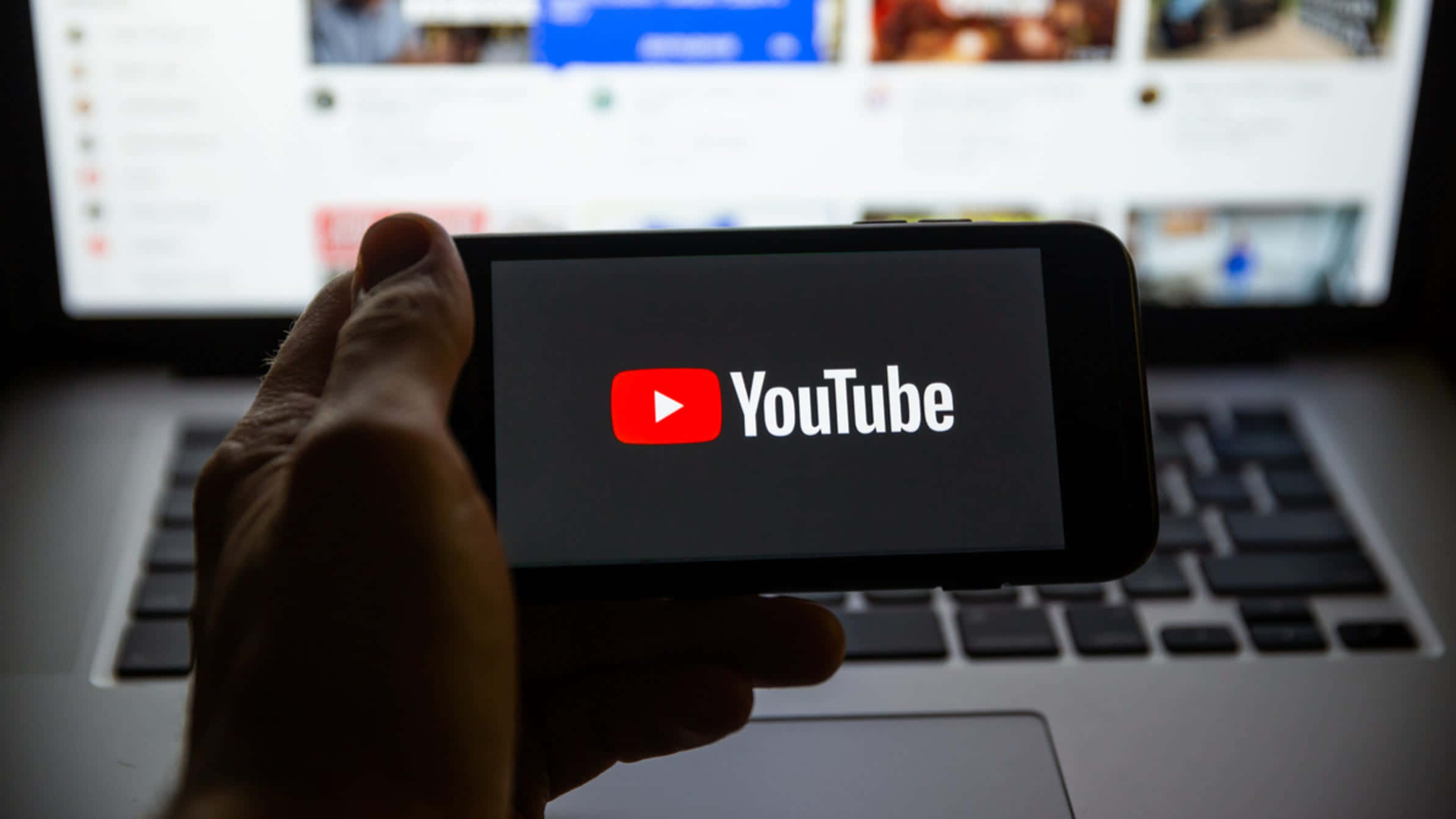 前所未有地體驗 YouTube 壁紙 — 一隻手拿著顯示YouTube標誌的智能手機，前面是一部顯示YouTube主頁的筆記本電腦。