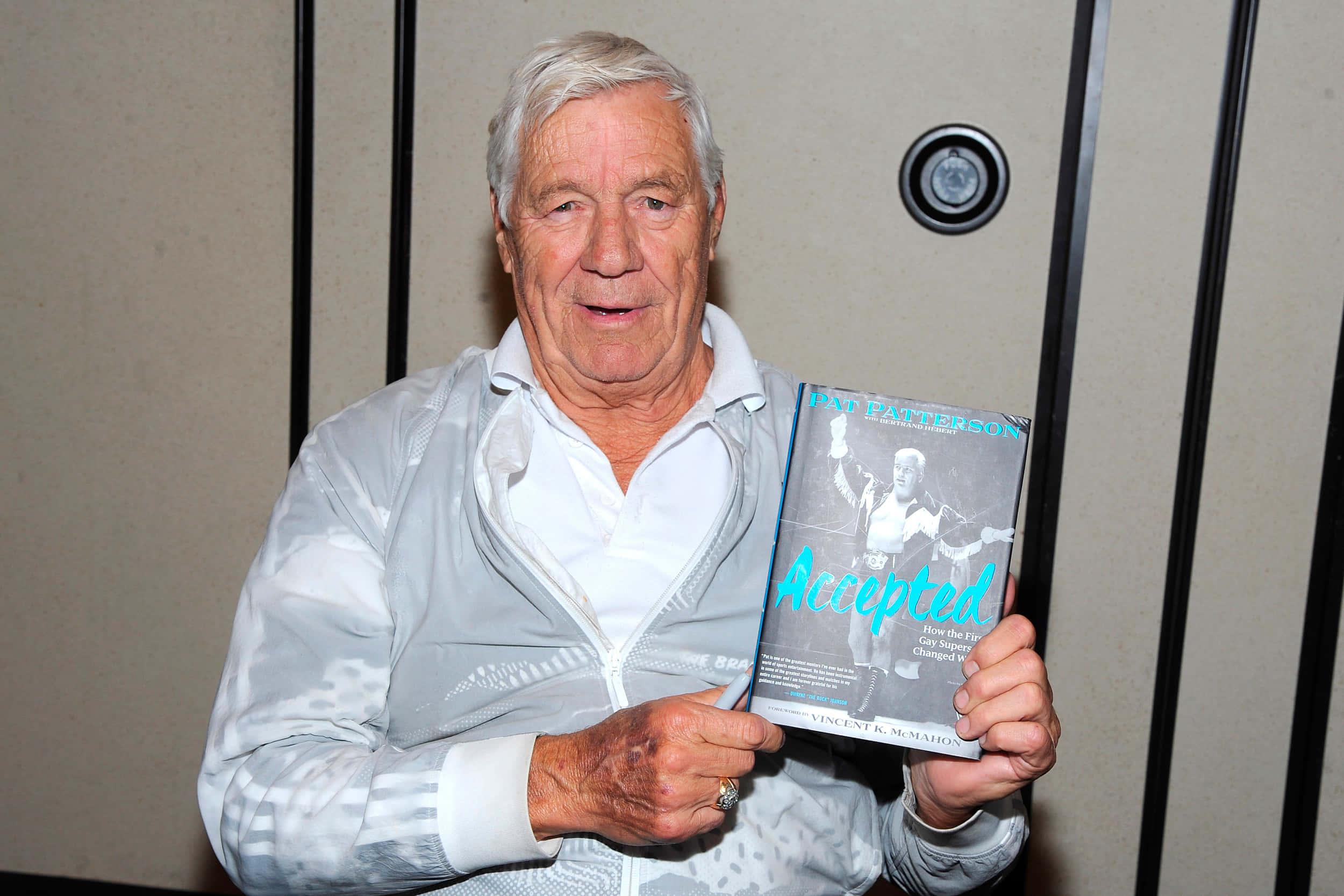 前摔角手 Pat Patterson 持書 壁紙 — 一位白髮老人在米色牆壁前微笑，牆上有黑色垂直條紋，他手持一本名為《Accepted》的書。