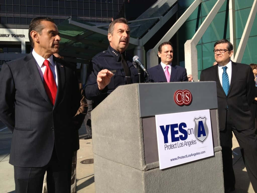 前洛杉磯市長安東尼奧·維拉雷加薩參與「Yes on Protect LA」運動 壁紙 — 四名身穿西裝和制服嘅男士企喺戶外講台上，帶住一個「YES on A Protect Los Angeles」嘅標語牌。