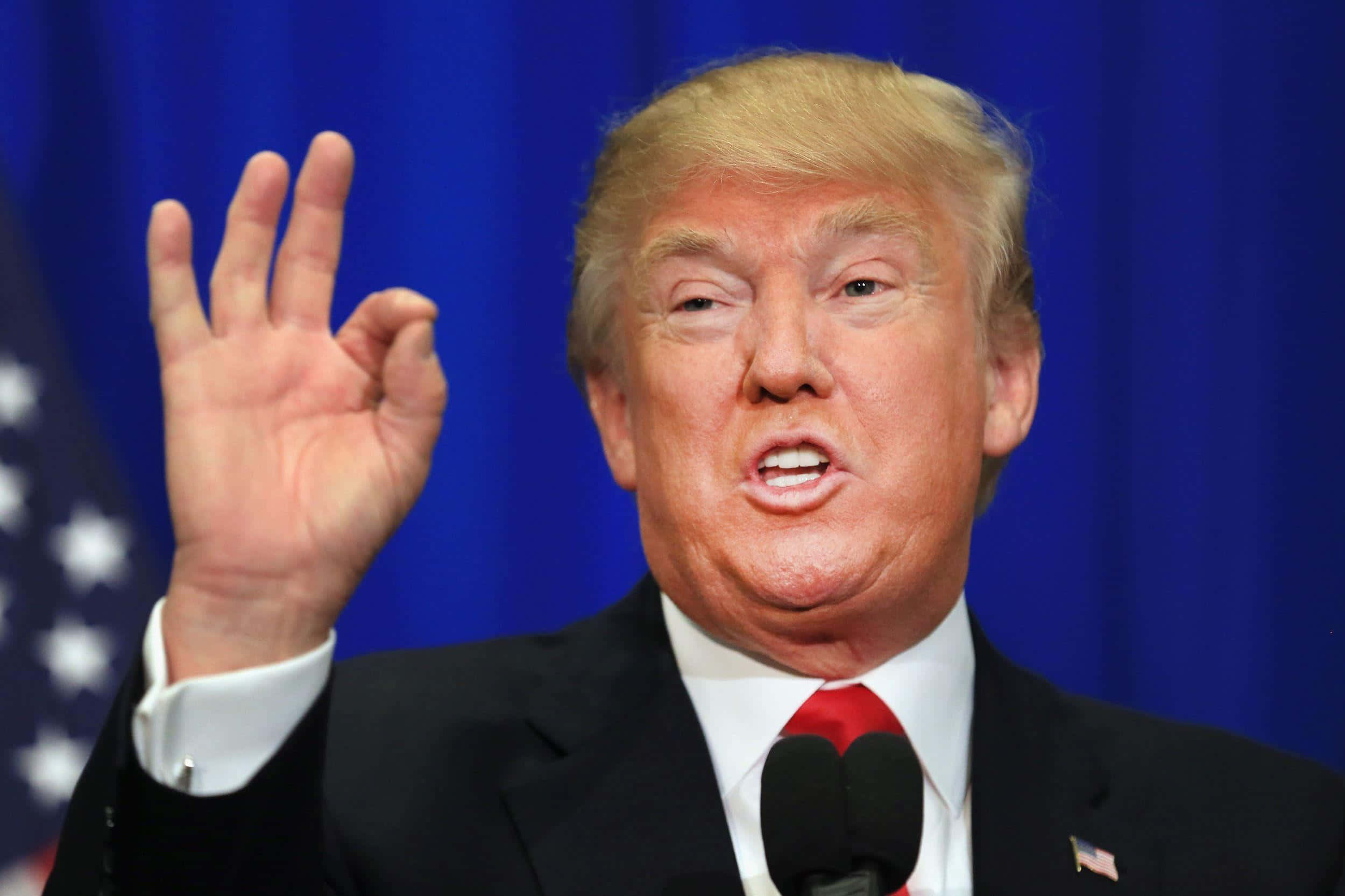 trump 桌布 壁紙 — 前總統唐納德·特朗普在美國國旗背景下，在講台上強調地做手勢發言。