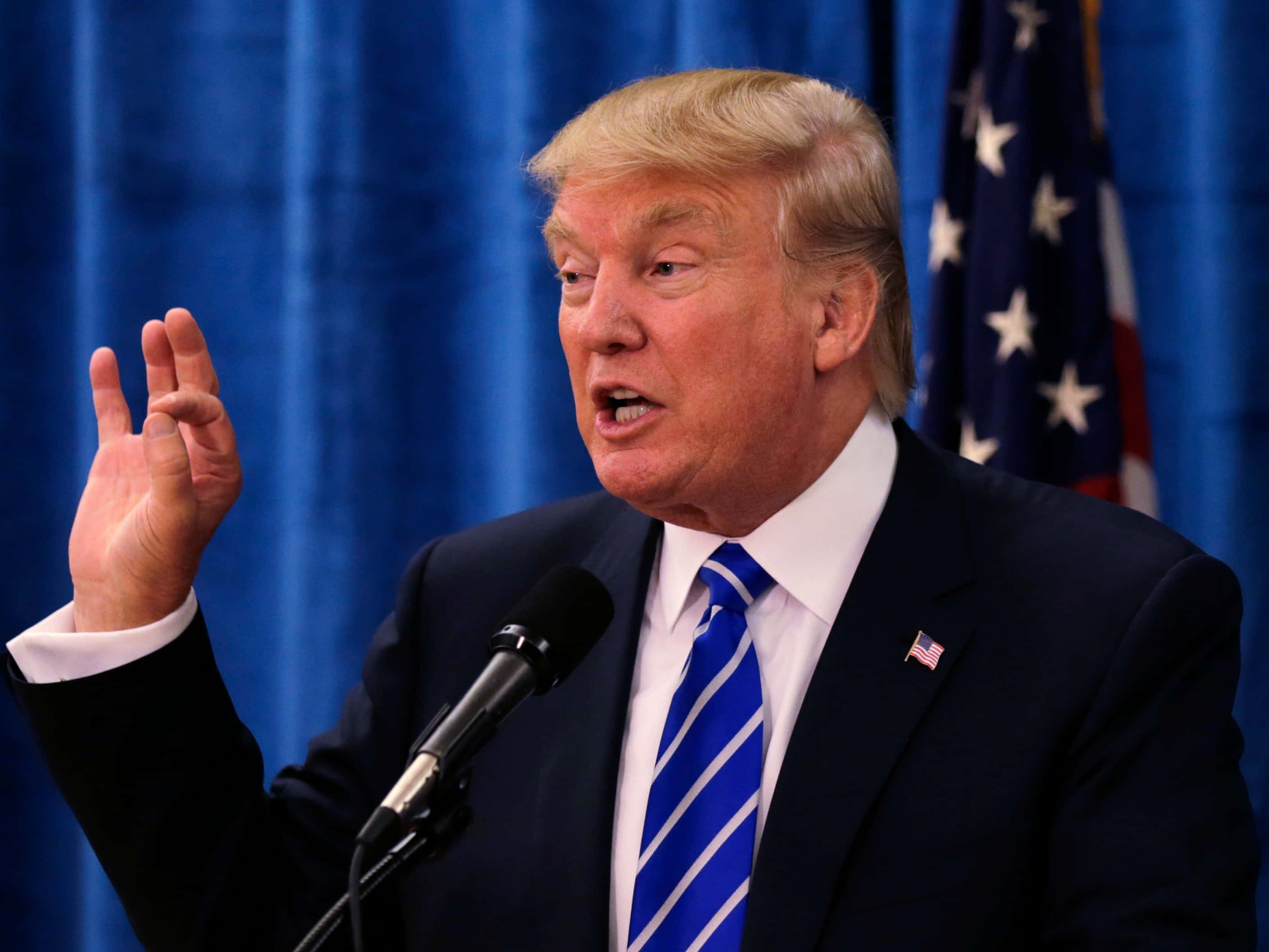 trump 桌布 壁紙 — 前總統當勞·特朗普在藍色帷幕和美國國旗背景下，對著麥克風說話時強調地做手勢。
