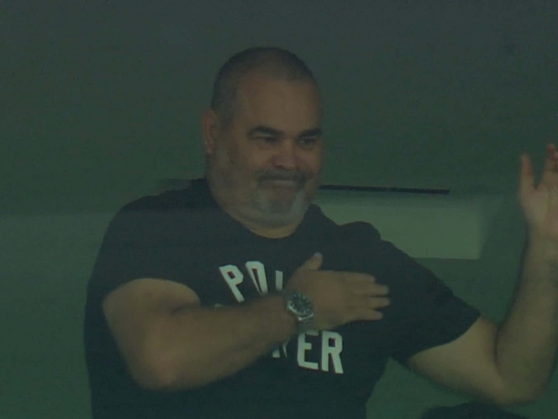 前足球員José Luis Chilavert於球場觀賽的隨拍 壁紙 — 一名身穿深色T恤的男子在燈光昏暗的房間裏，雙手比劃著，露出溫暖的笑容。