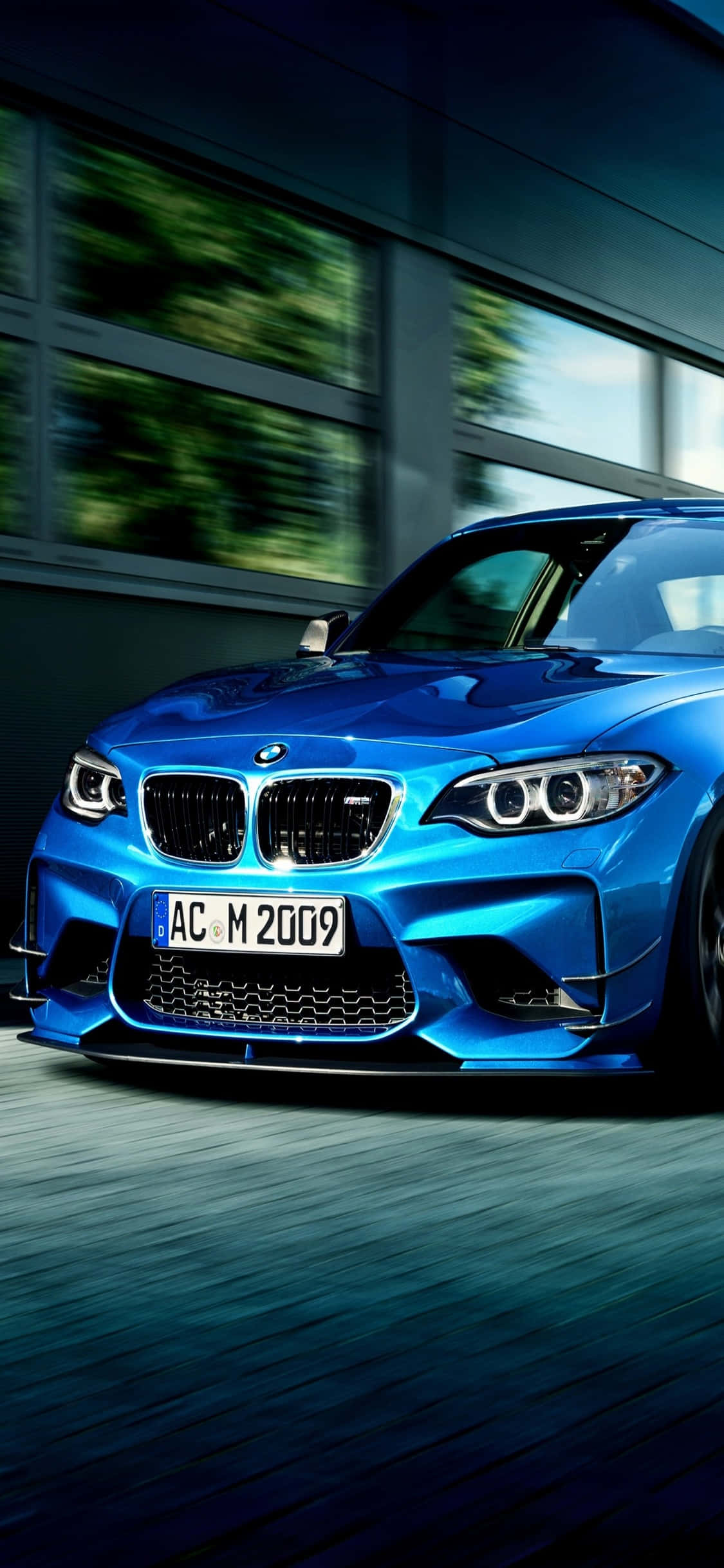 剛下線的時尚組合 — iPhone X 與 BMW 壁紙 — 一輛鮮藍色BMW M2跑車以動態模糊效果高速駛過一棟現代建築，強調其動感。