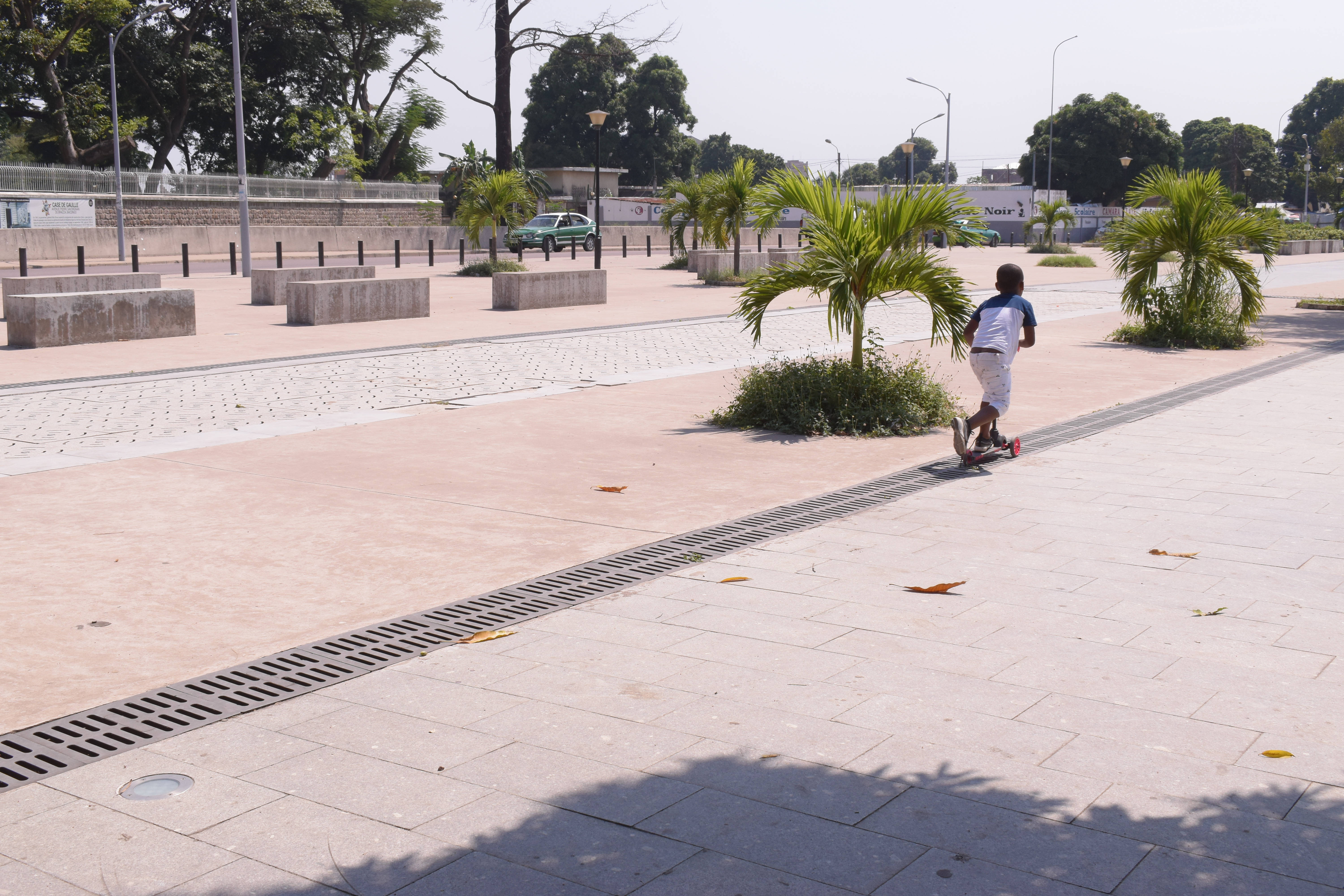 剛果晴朗街道 壁紙 — 一個小孩在陽光普照的鋪砌廣場上騎滑板車，廣場上有棕櫚樹和混凝土長椅。