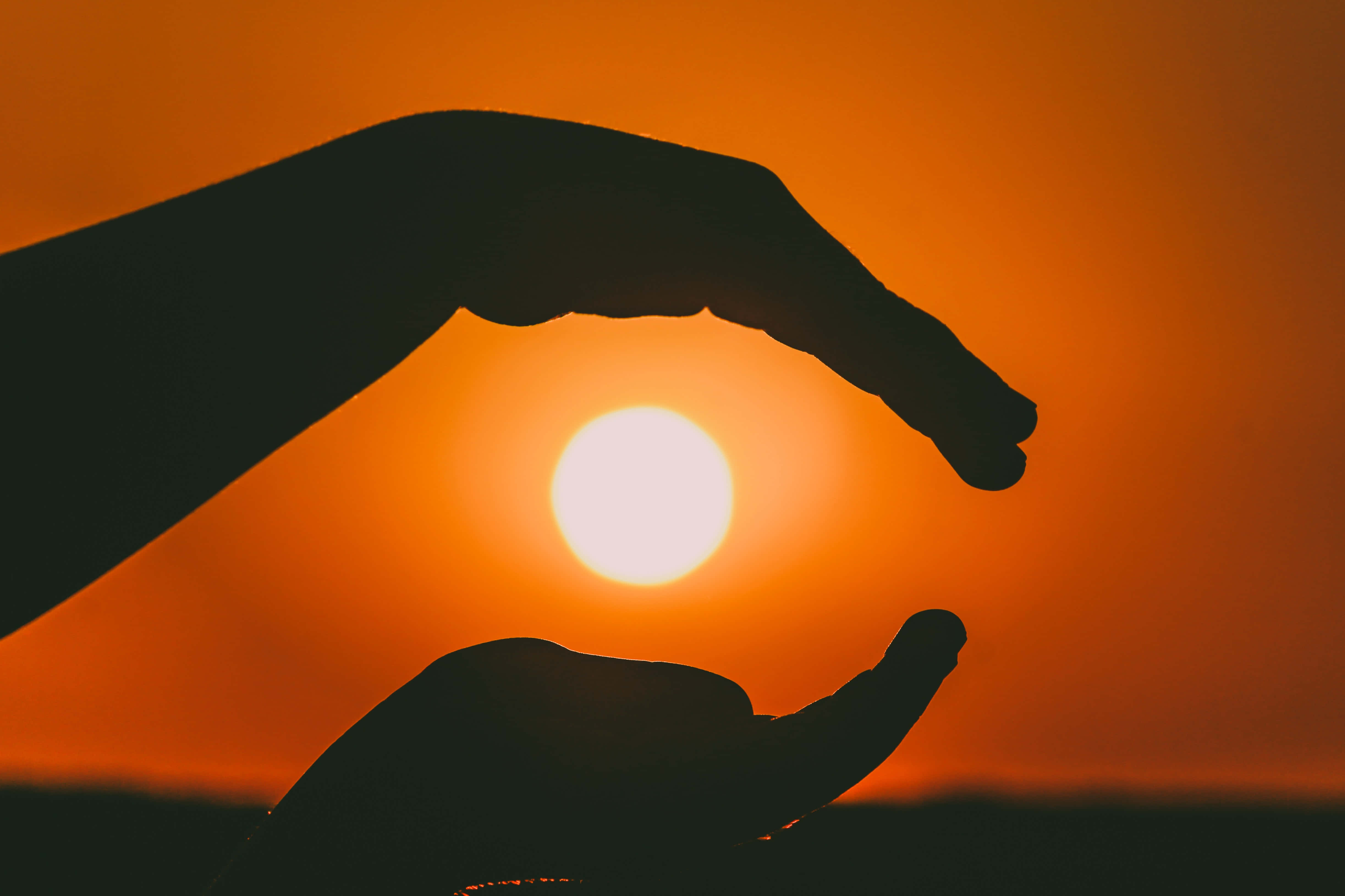 hands 桌布 壁紙 — 剪影中，雙手輕輕捧著一顆發光的太陽，背景是日落時溫暖的橙色天空。