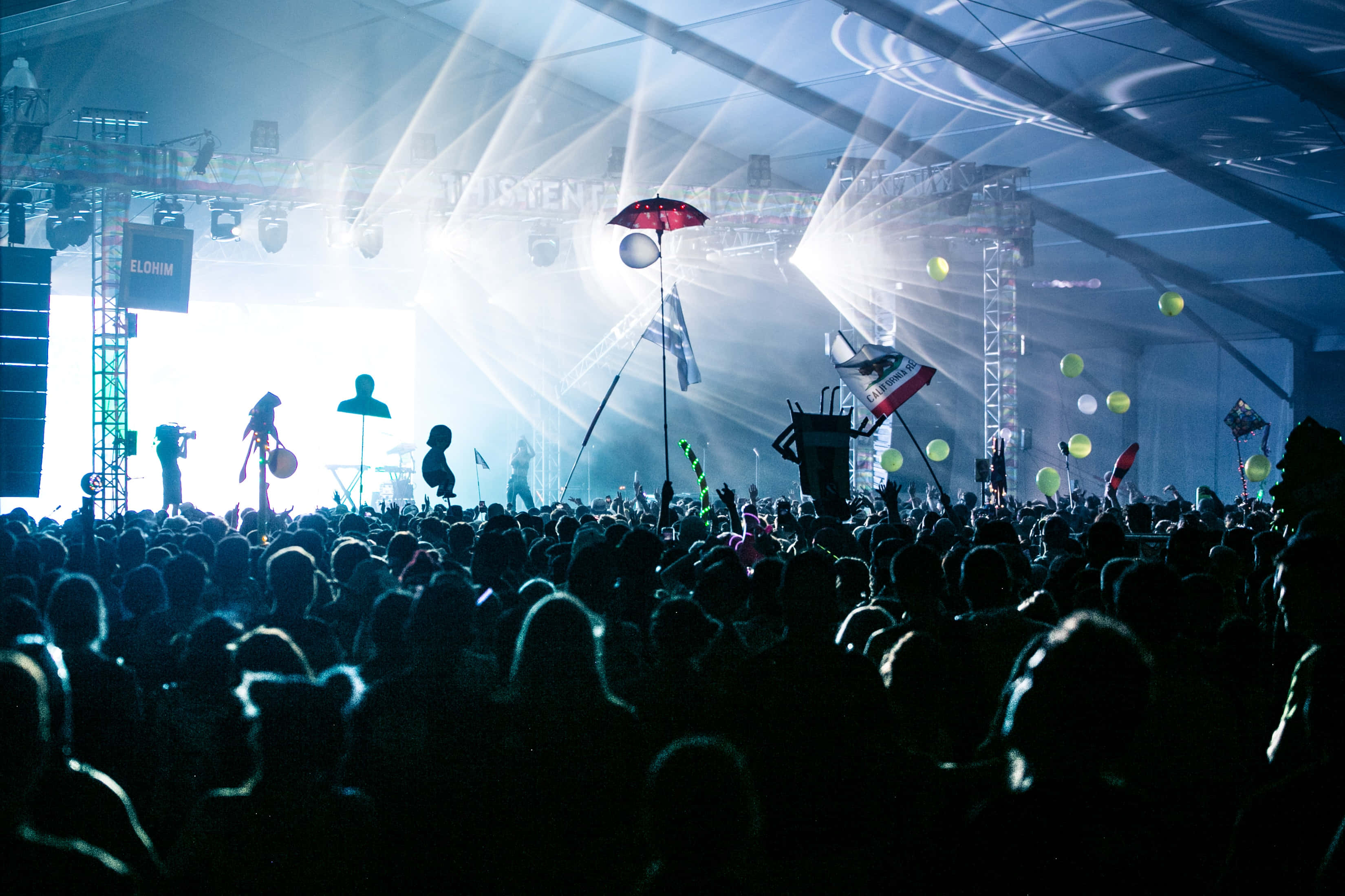 music festival 桌布 壁紙 — 剪影人群在明亮的舞台燈光下，於一個有漂浮氣球和旗幟的大型室內演唱會中向前湧動。