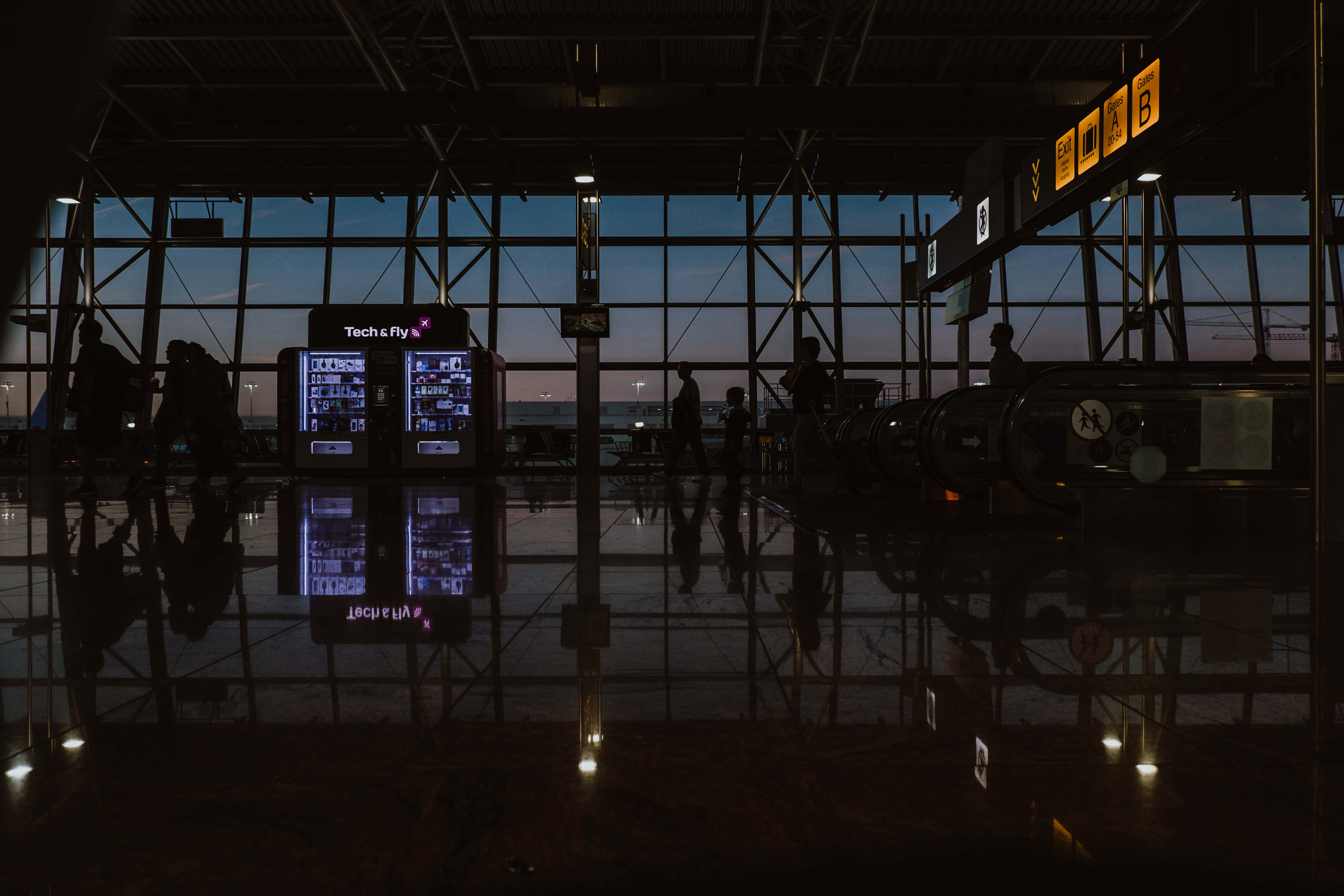 Airport 桌布 壁紙 — 剪影旅客在黃昏時分穿過昏暗的機場客運大樓，周圍有發光的屏幕和反光地板。
