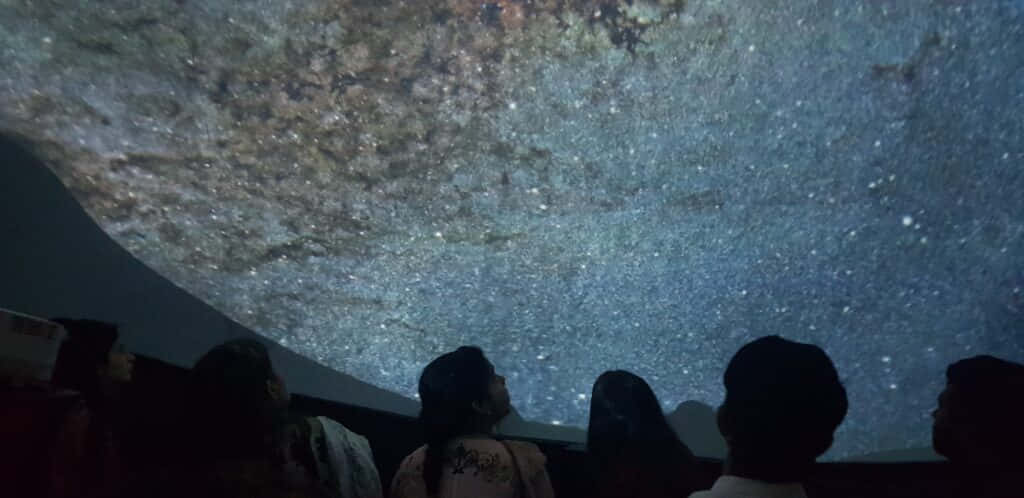 Planetarium 桌布 壁紙 — 剪影般的觀眾敬畏地仰望一個廣闊的沉浸式圓頂投影，呈現出繁星點點的夜空和發光的星雲。