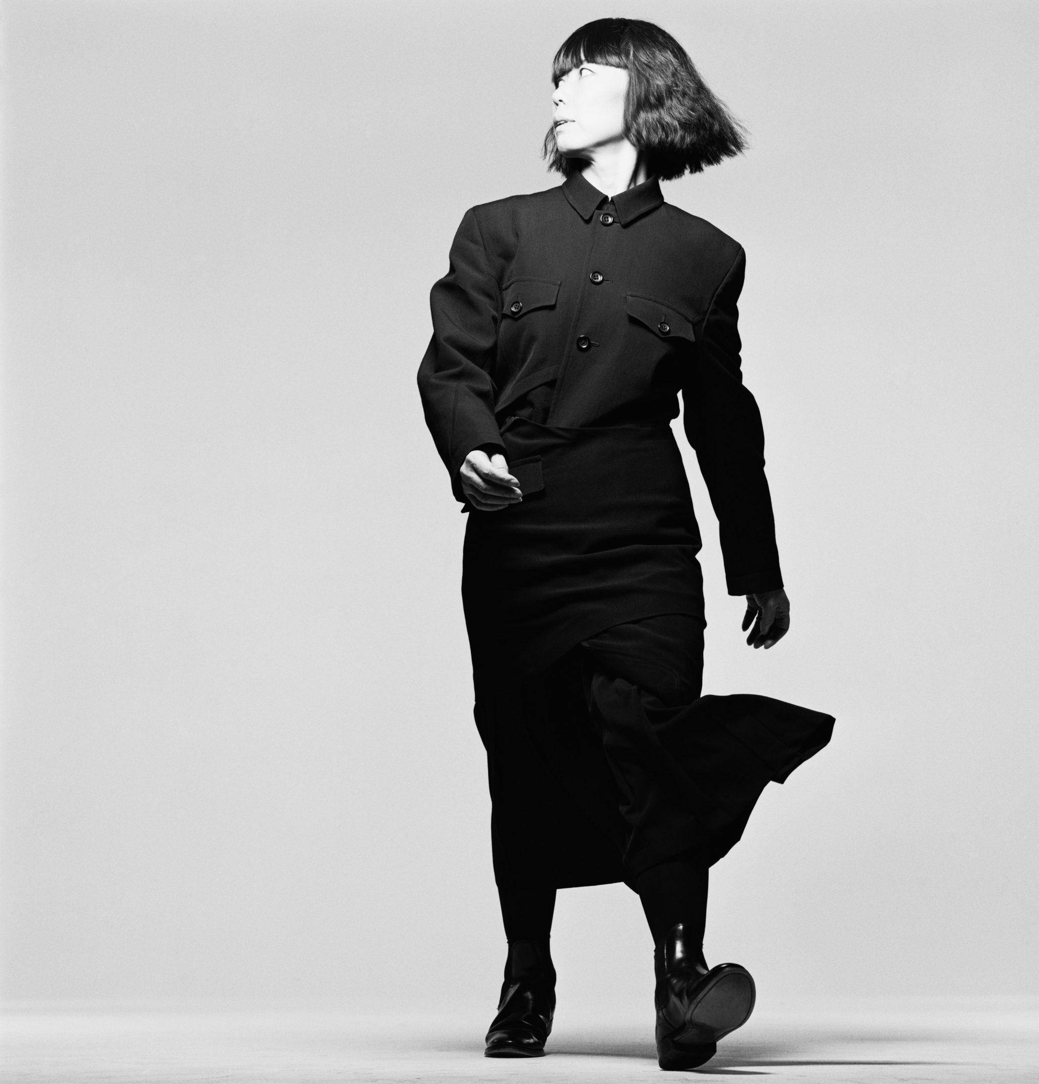 創意設計師 Rei Kawakubo —— Comme des Garçons 的創始人 壁紙 — 一位身穿黑色結構連衣裙的女性在單色調的純灰色工作室背景下自信地大步走。