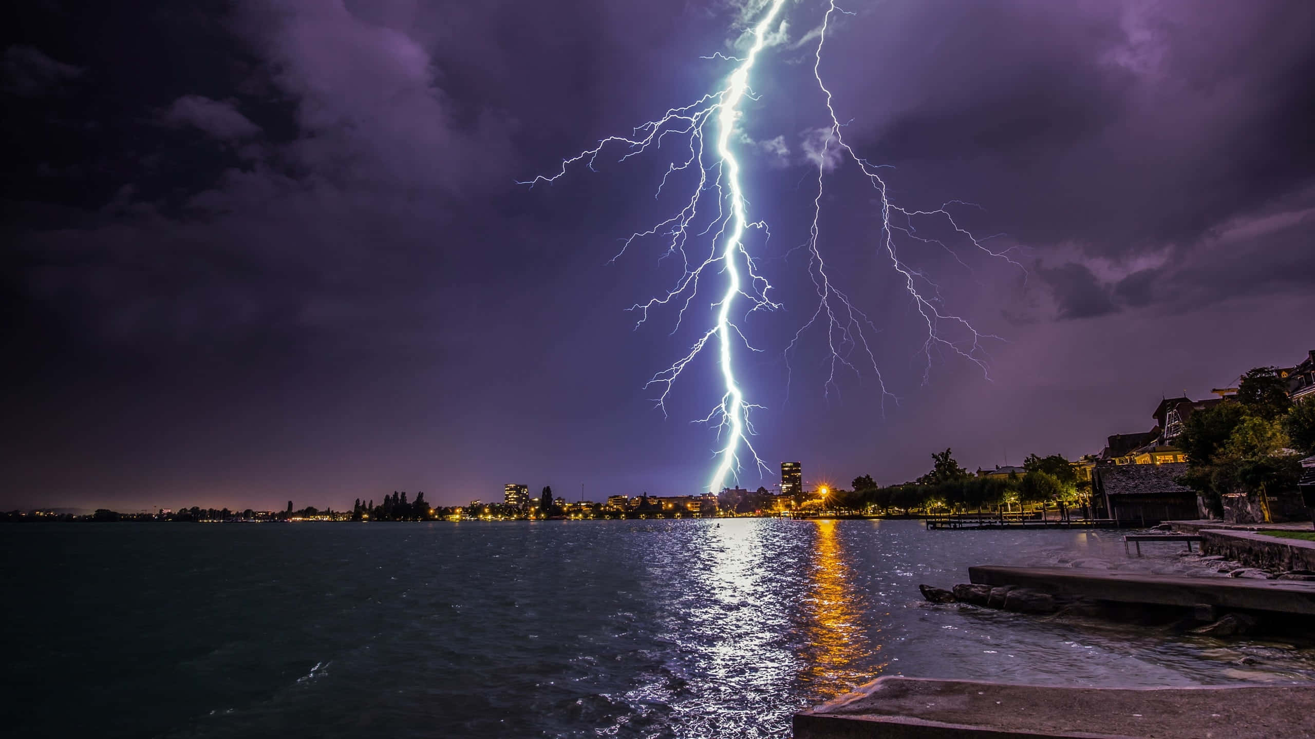 劇烈的暴風雨天空 壁紙 — 一道巨大的閃電在夜間劃過黑暗的湖面，照亮了湖水與遠處的城市燈光。