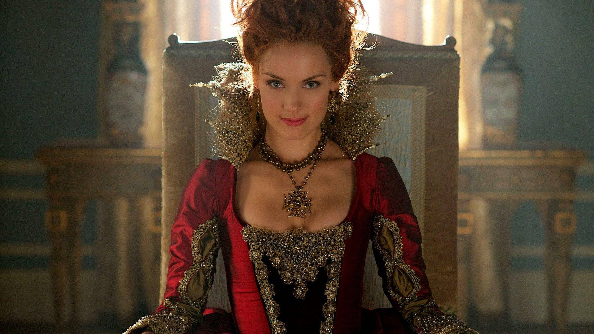 劇集《Reign》凱瑟琳·德·美第奇女王 壁紙 — 一位身穿紅金色文藝復興長袍的尊貴女性，端莊地坐在陽光普照的華麗王座上。