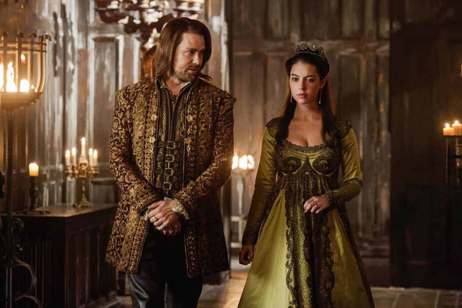 劇集《Reign》塞巴斯蒂安王子與瑪麗女王 壁紙 — 一名身穿華麗中世紀服裝的男子和女子，在燭光照亮的石砌大廳中，穿過厚重的木門和吊燈。