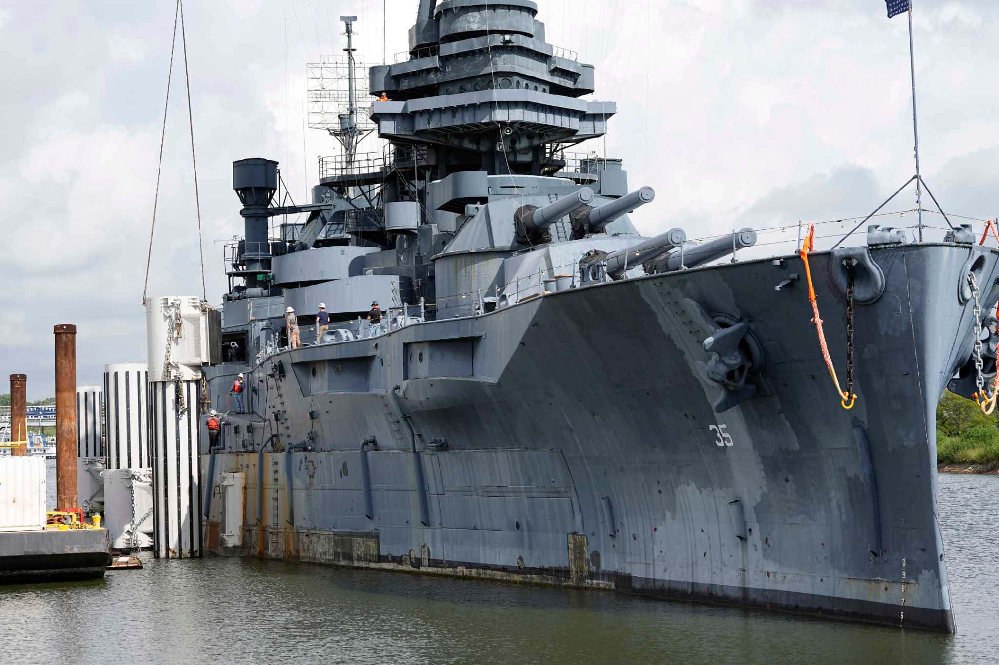 劈波斬浪：戰艦緩緩靠岸 壁紙 — 一艘巨大的灰色戰艦，帶有突出的炮塔和甲板炮，停泊在碼頭旁的平靜水域，天空多雲。