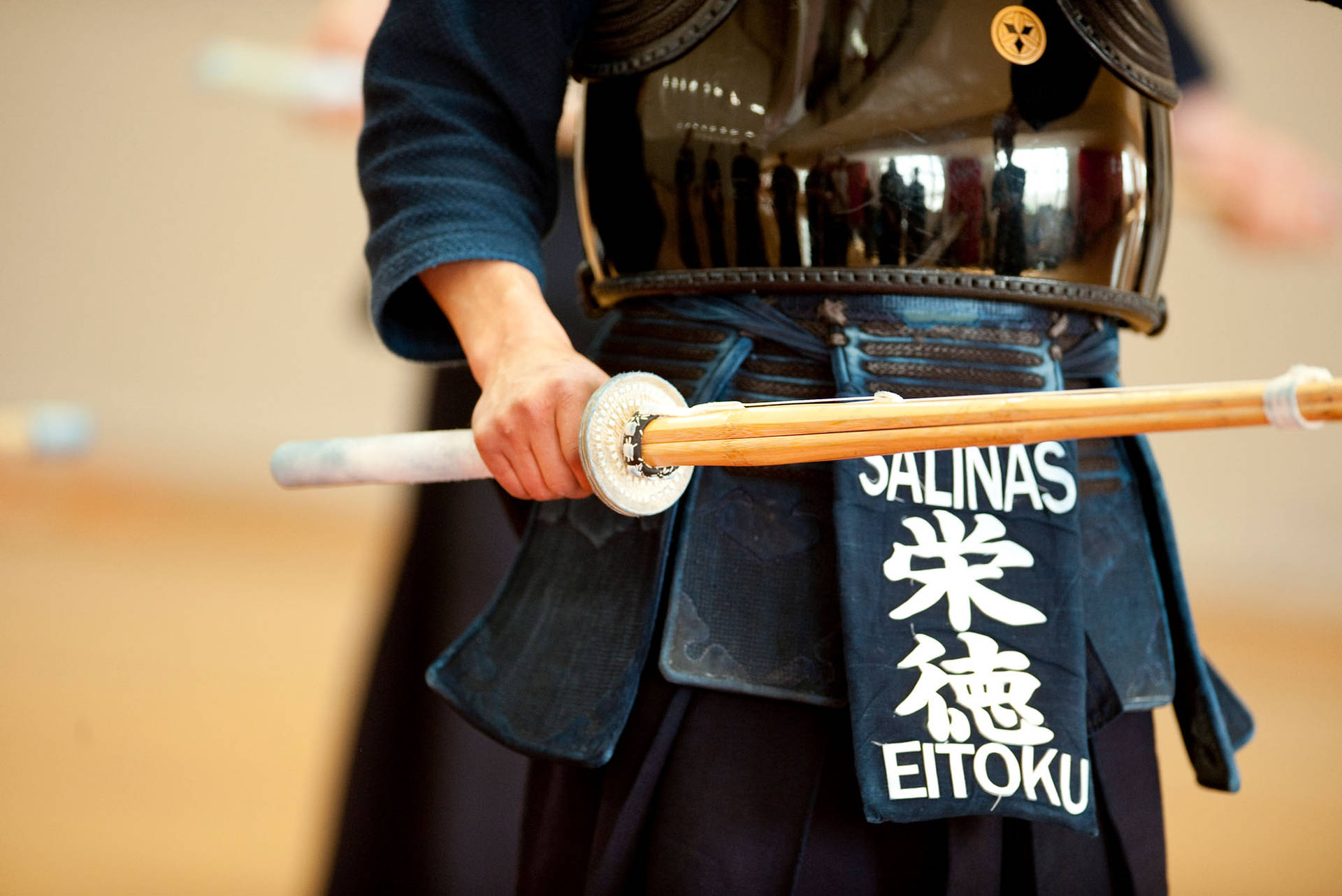 劍道練習者握持竹劍 壁紙 — 一位身穿黑色盔甲的武士，在模糊的室內背景下握著一把白色刀柄的木劍。