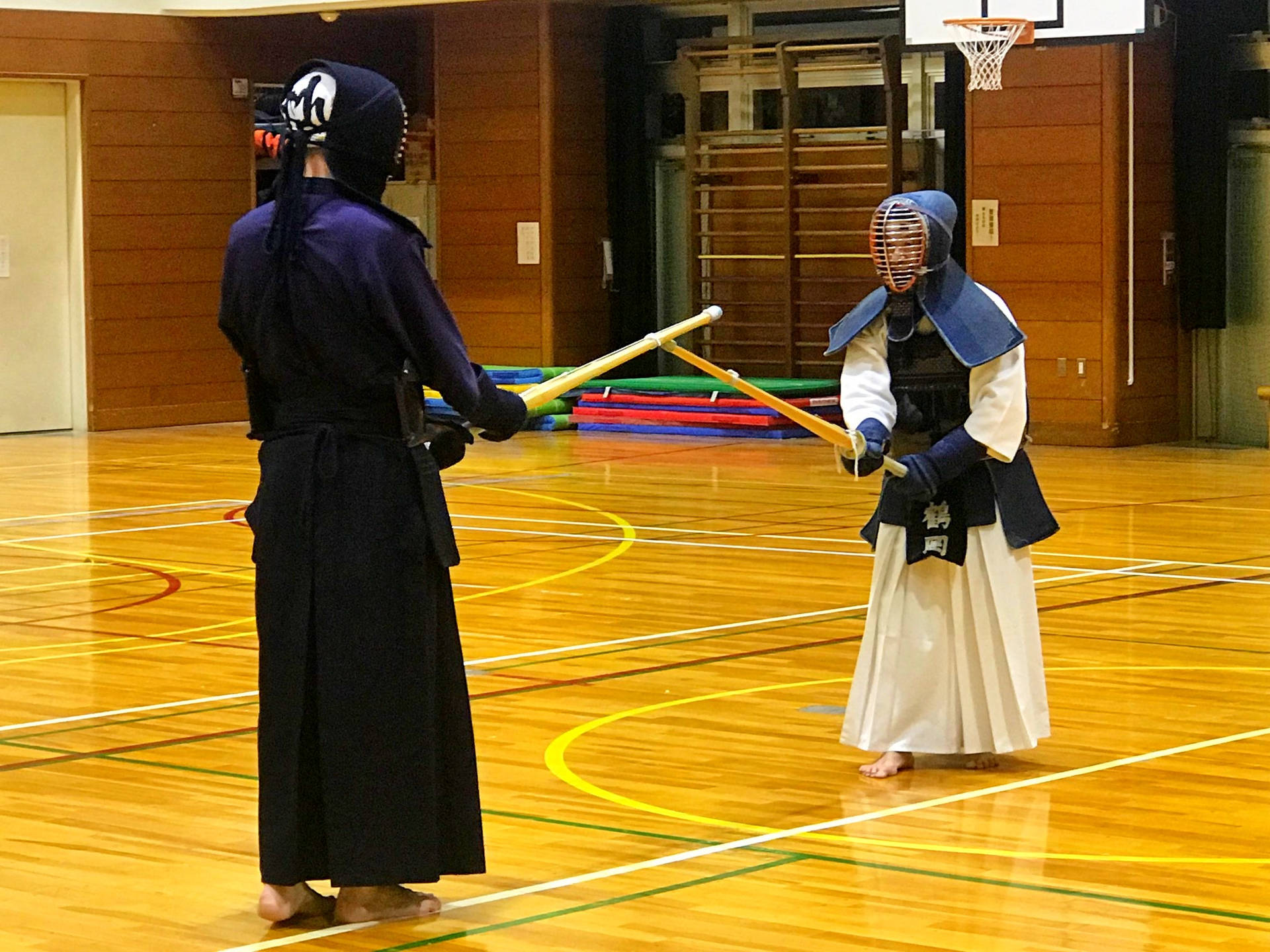 劍道選手Kenji Inukai與Tsuruoka老師對決 壁紙 — 兩名全副裝備的劍道練習者在明亮的室內燈光下，於拋光木質體育館地板上交劍。