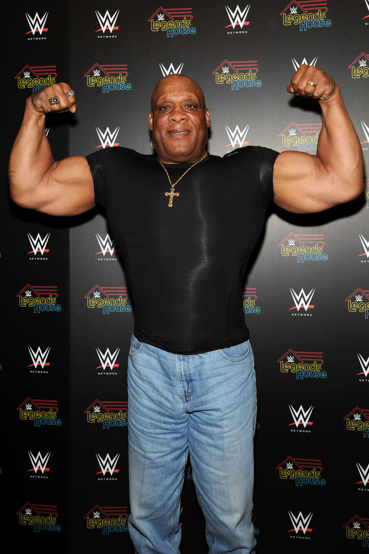 WWE摔角手在Legends House活動中彎曲二頭肌，穿著黑色襯衫和牛仔褲。