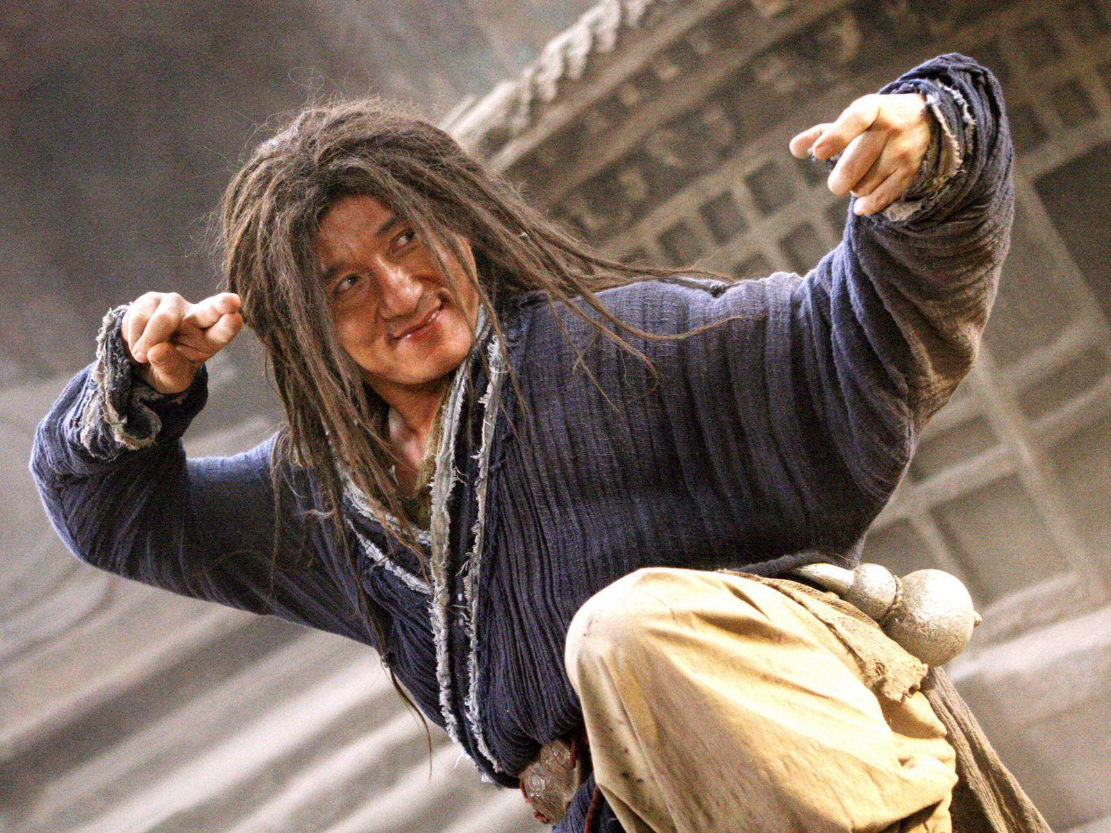 《功夫之王》成龍 壁紙 — 一個頭髮凌亂、咧嘴而笑的男人，在模糊的石建築背景下擺出動態的武術姿勢。