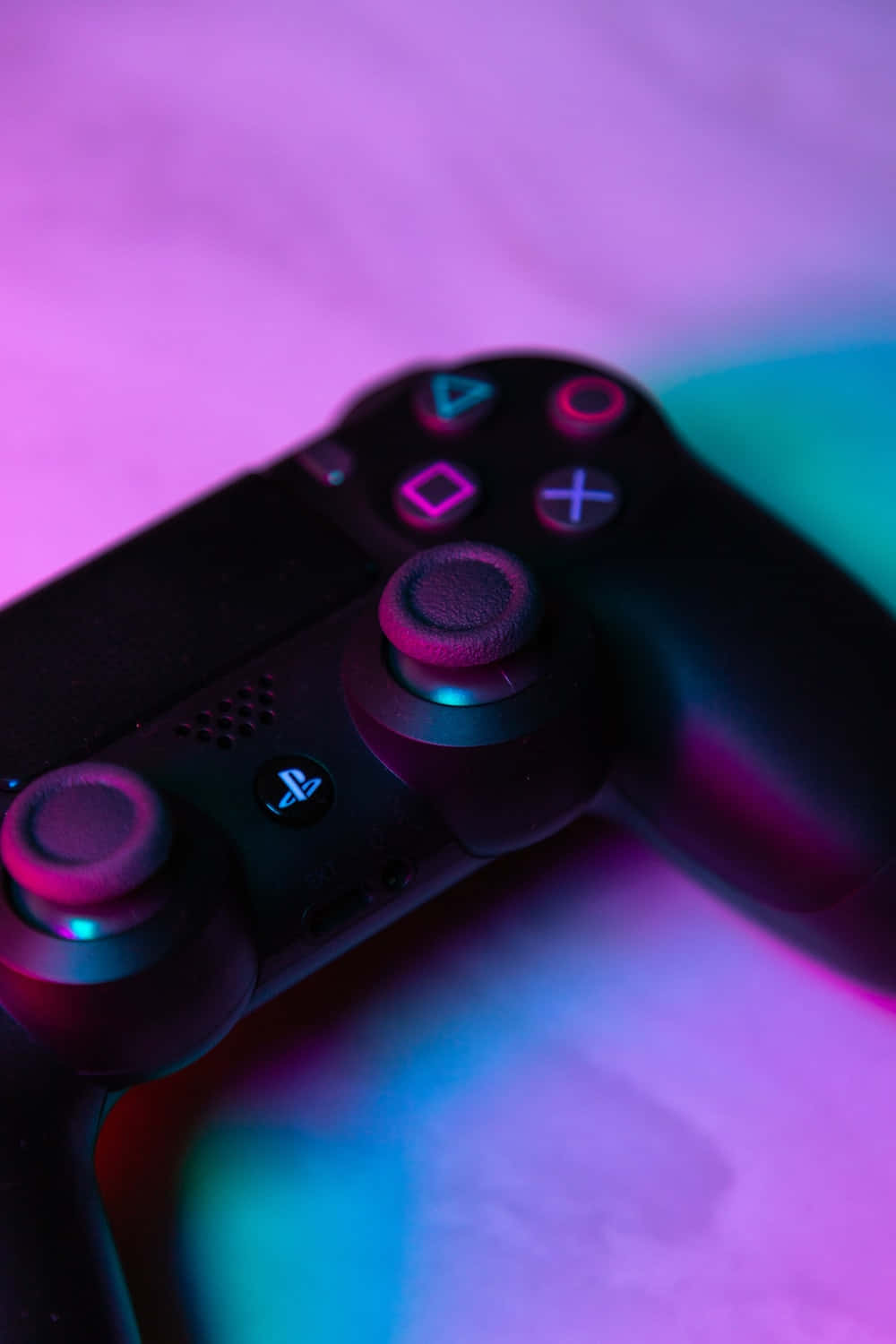 加入下一個遊戲等級：PlayStation 壁紙 — 黑色PlayStation手掣喺柔軟表面上被鮮豔嘅粉紅同青藍色霓虹燈照射嘅特寫。